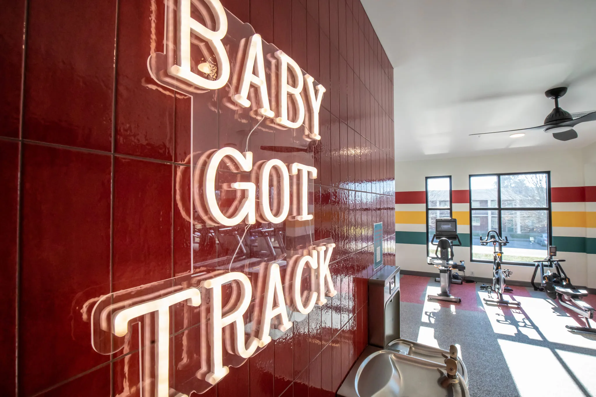Student housing amenity gym entrance: neon sign reading Baby Got Track glowing on oxblood tile