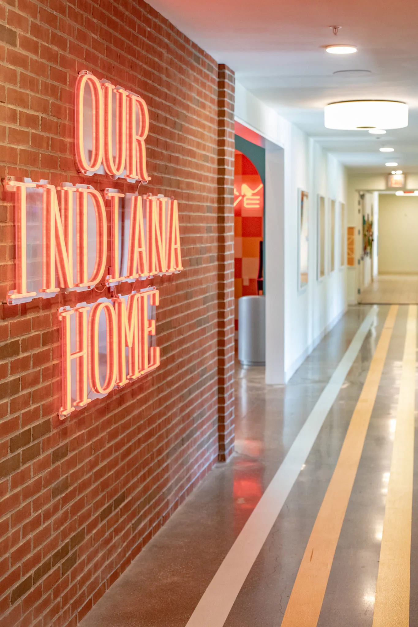 Student housing corridor with a neon sign reading Our Indiana Home on exposed brick beside striped track lines in the floor