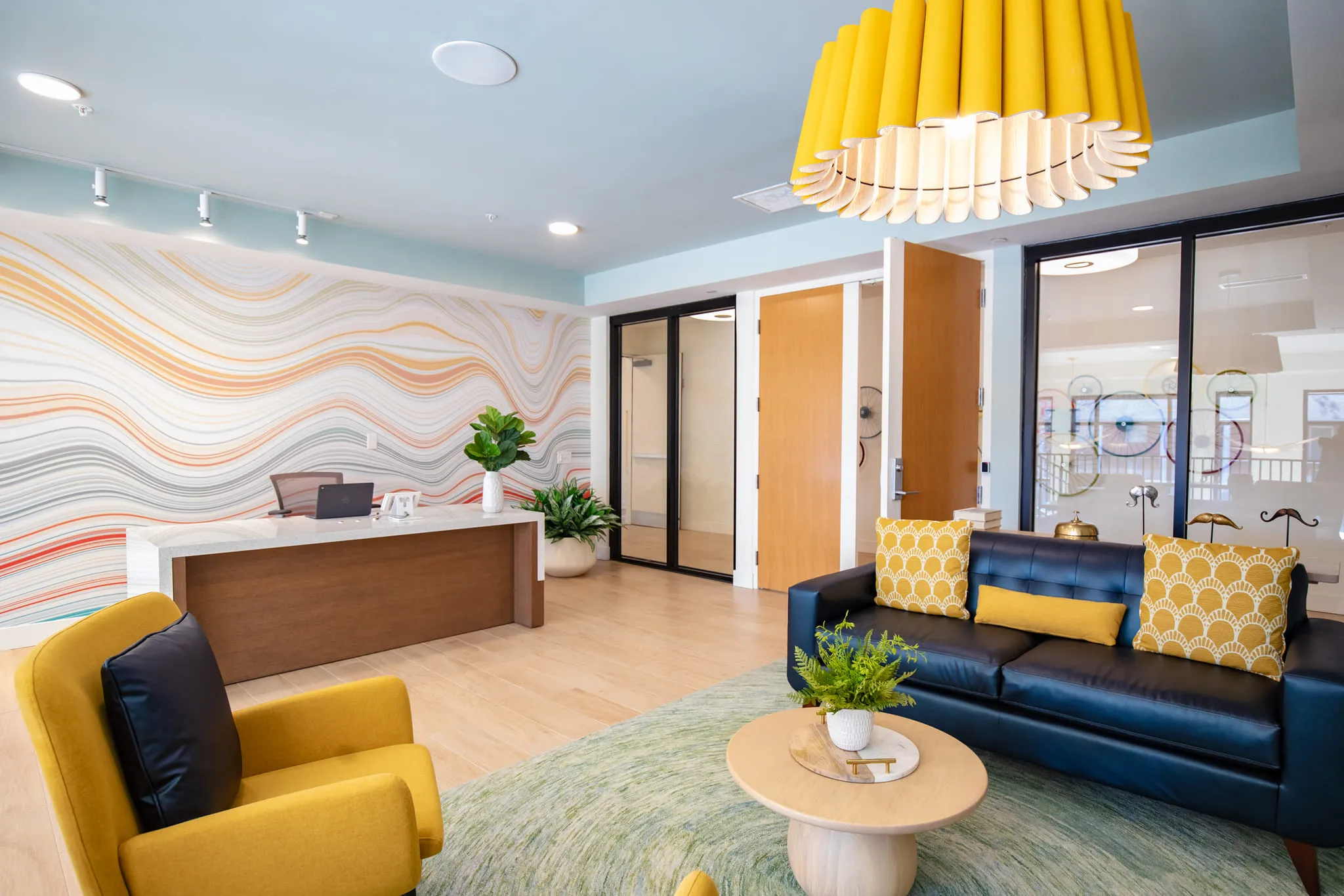 Student housing amenity leasing office with a navy sofa and yellow chairs on a green rug before a layered wave mural