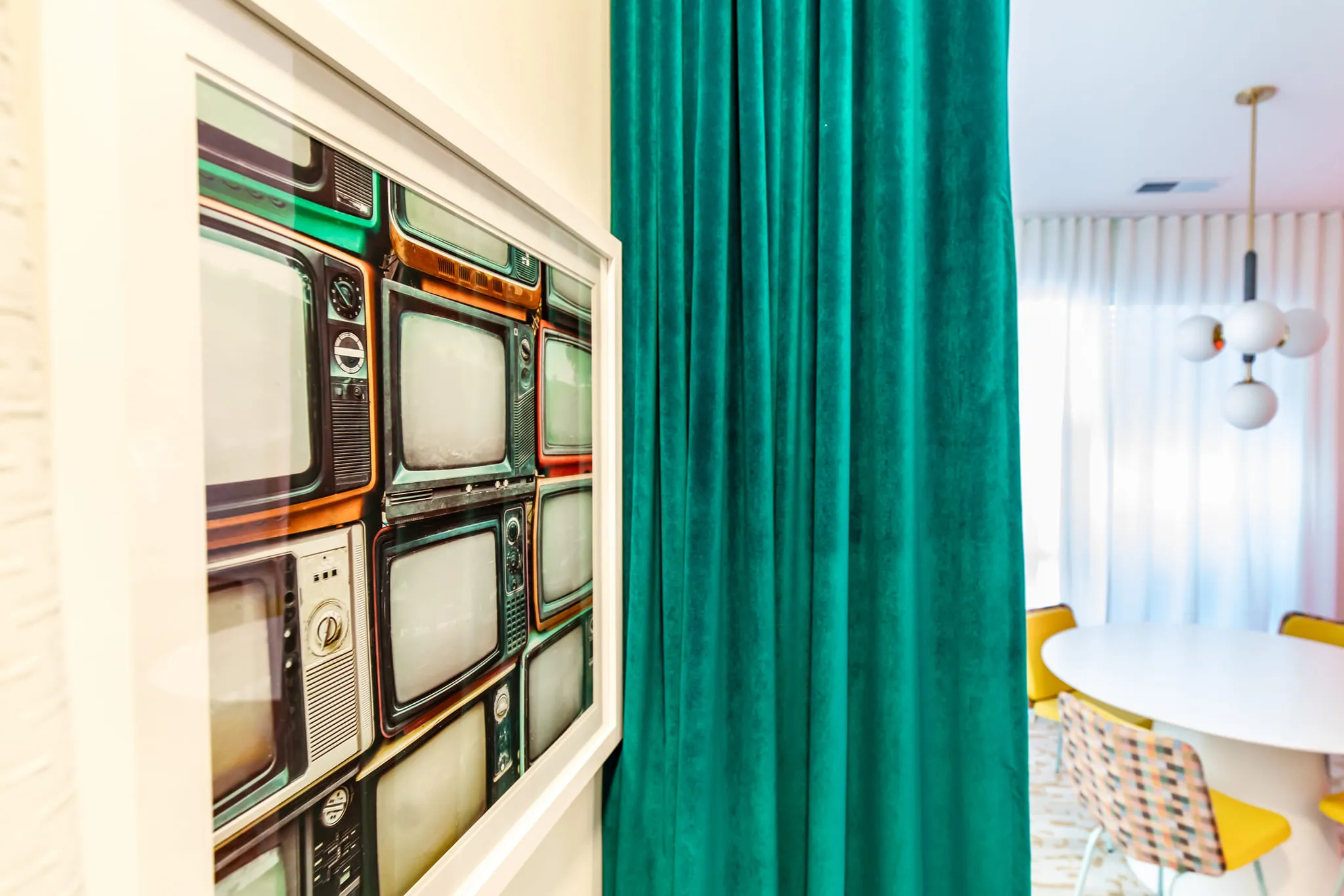 Student housing interior detail: framed print of stacked vintage TVs beside a teal velvet curtain, yellow chairs beyond