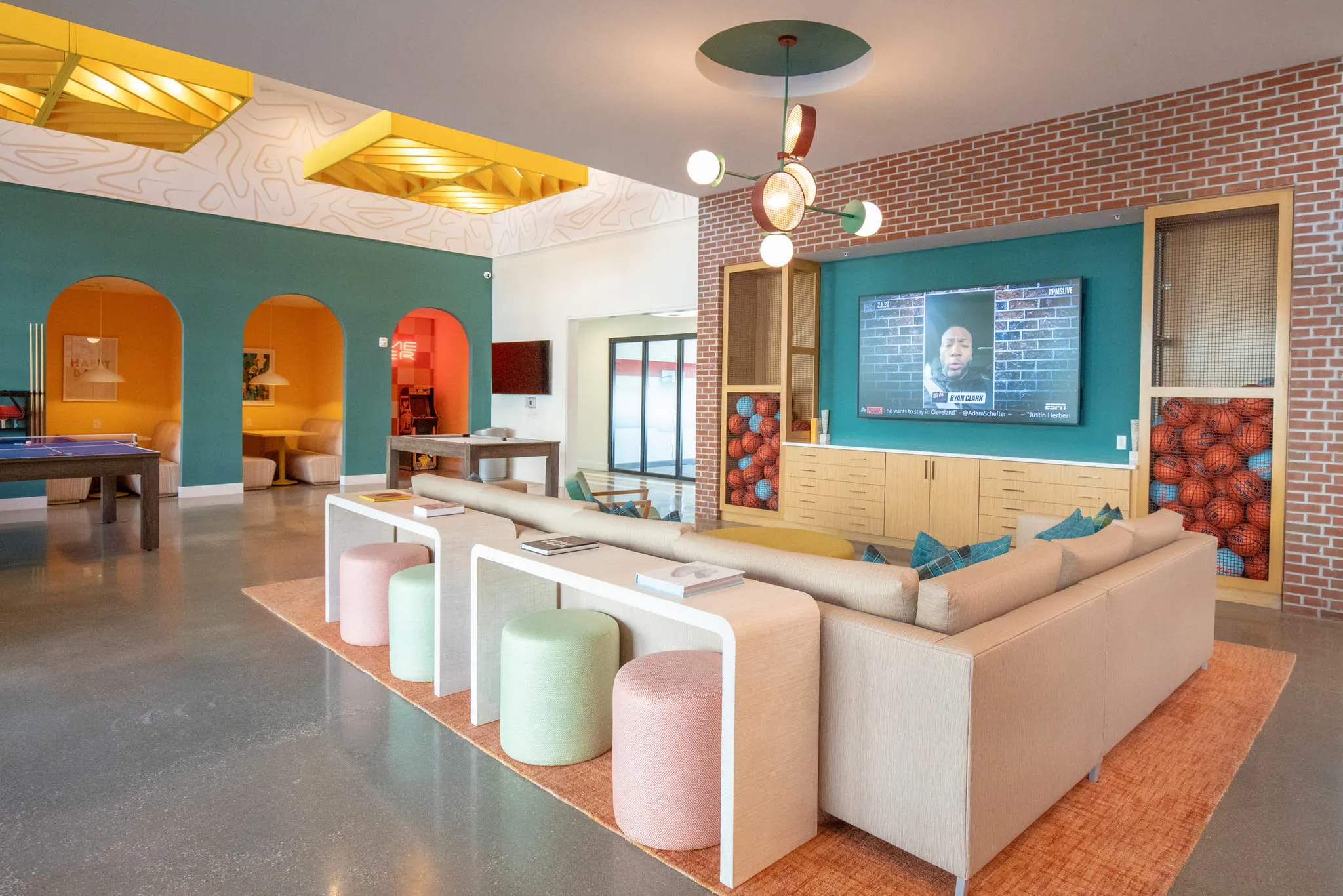 Student housing amenity sports lounge with a sectional and pastel ottomans facing a screen in cabinetry full of basketballs