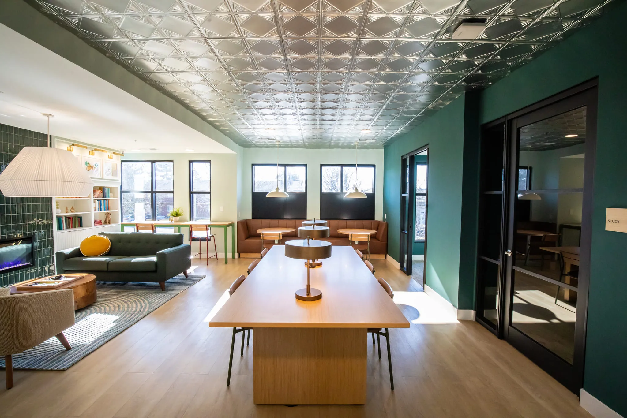 Student housing amenity study lounge with a long communal table under a pressed tin ceiling, glass study rooms alongside