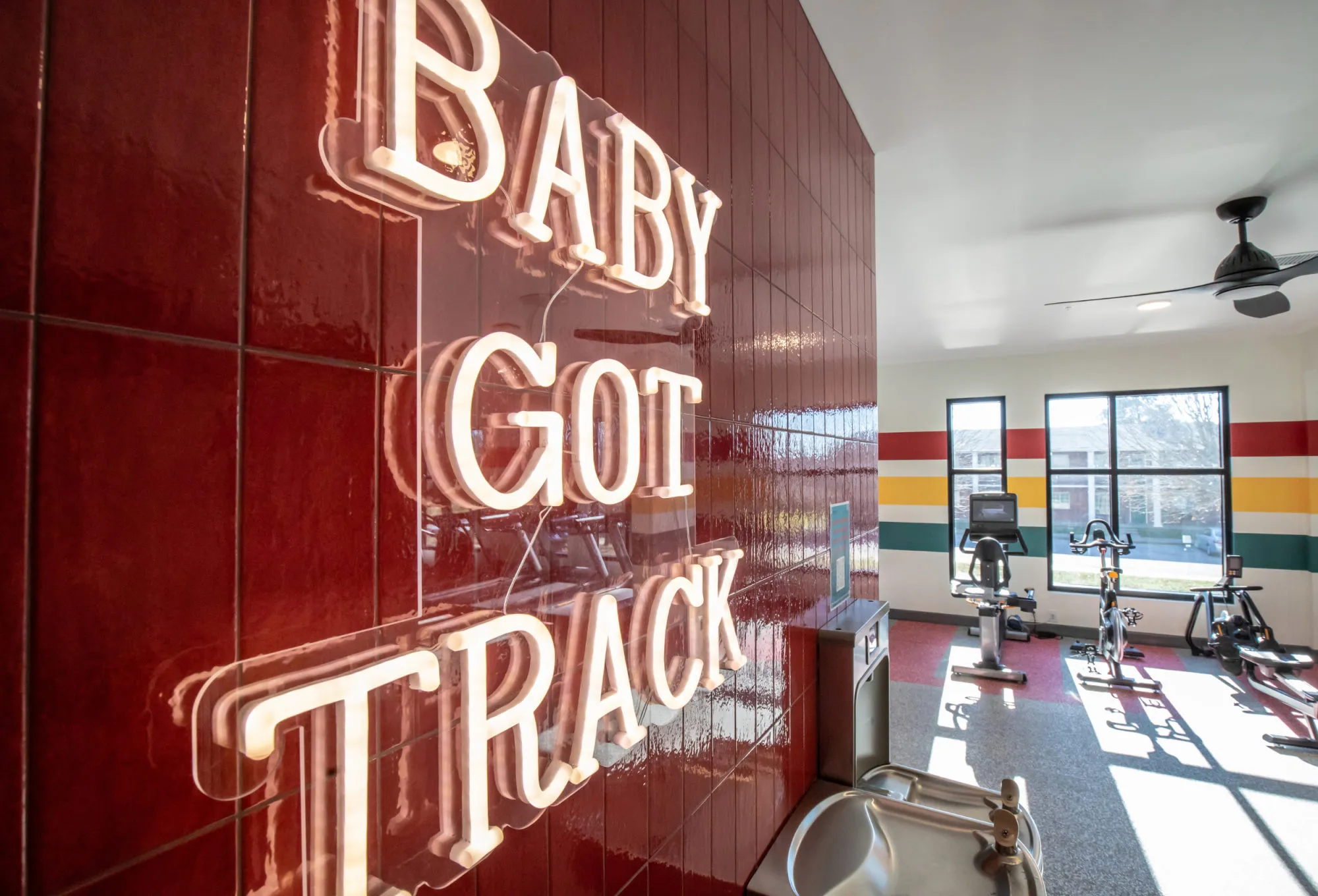 Student housing amenity fitness room with a neon Baby Got Track sign on red tile and spin bikes by a striped wall