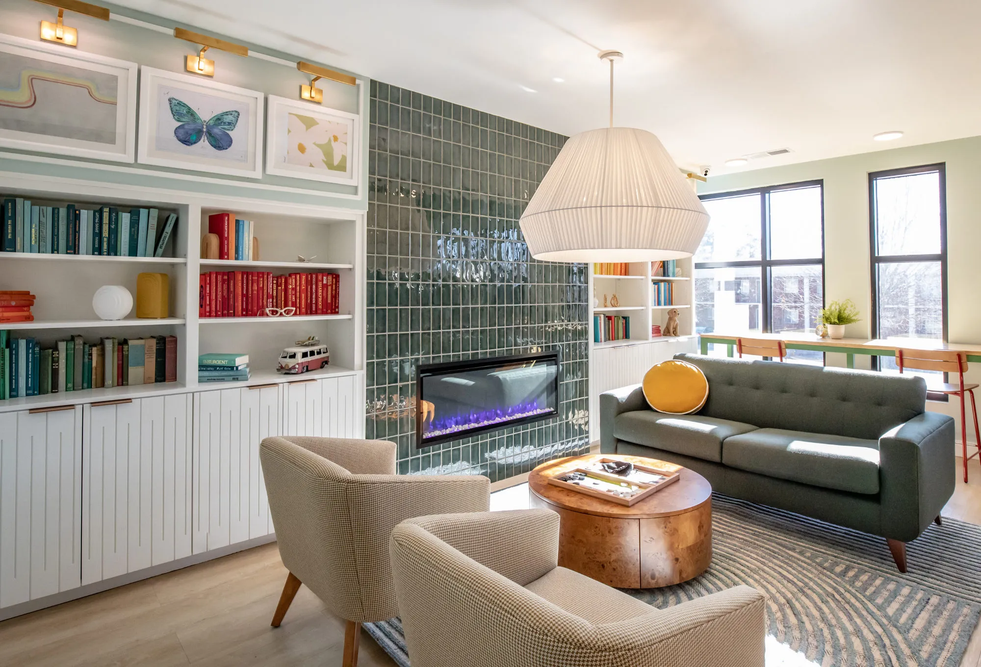 Student housing amenity study lounge with colour-sorted bookcases, butterfly prints, a green tile fireplace and a sage sofa