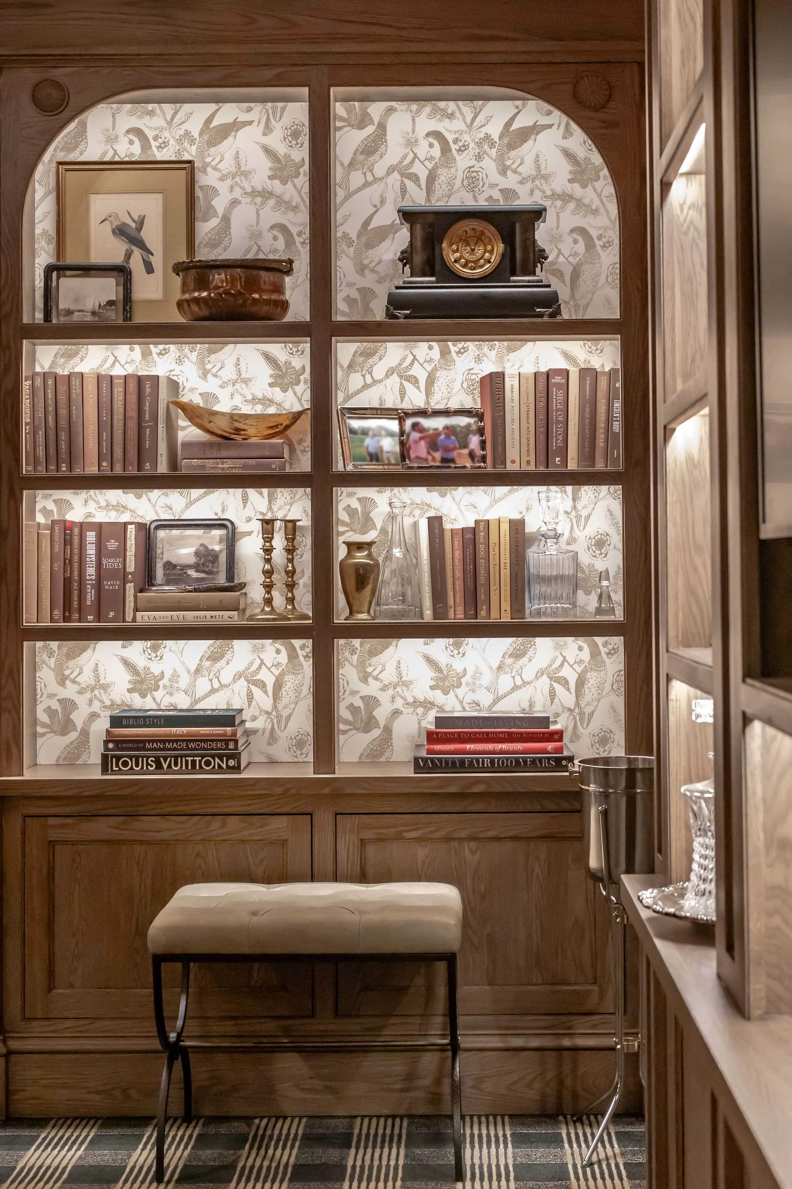 Built-in bookcase with patterned wallpaper behind the shelves, books and framed pictures under brass picture lights, and an upholstered stool in front