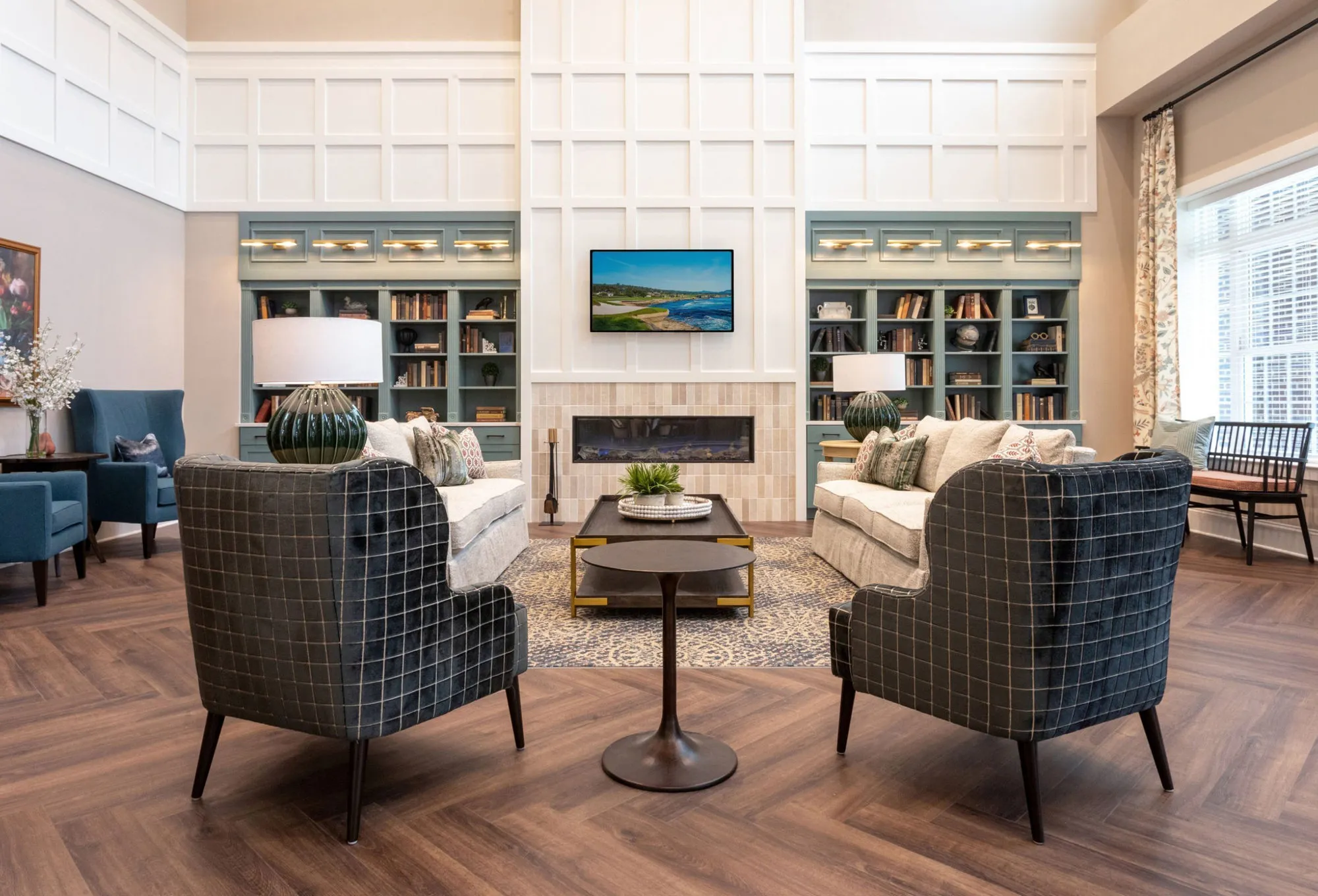 Two-storey lounge with a white panelled fireplace between teal bookcases and navy windowpane check wing chairs on herringbone