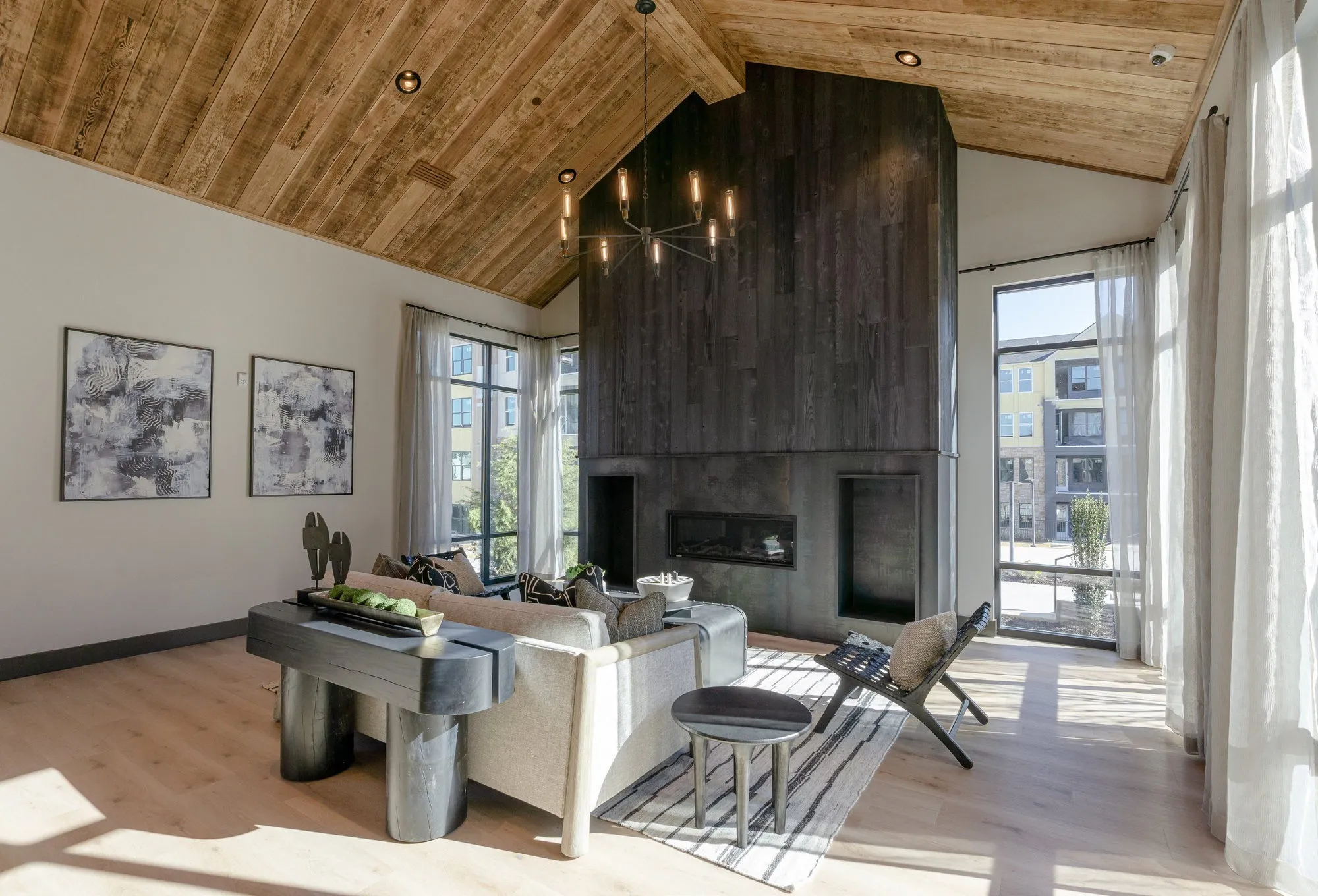 Clubroom under a vaulted wood ceiling with a blackened wood fireplace wall, a cream sectional and sheer-curtained windows