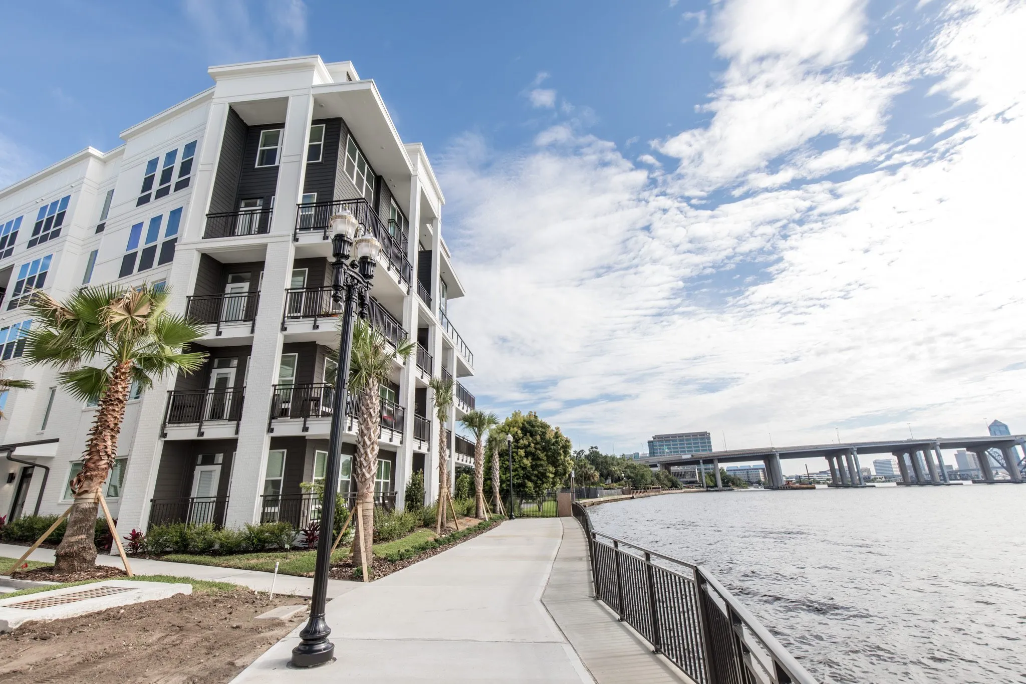 Multifamily community exterior: riverwalk with a black lamp post and railing beside the building, a bridge beyond