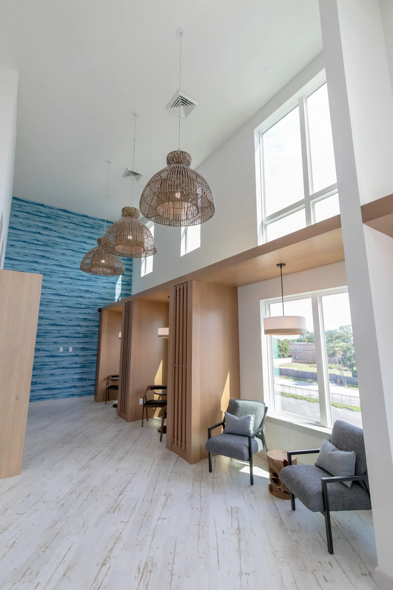 Multifamily amenity clubroom with grey armchairs by a window, booths, woven pendants and the blue water pattern wall beyond
