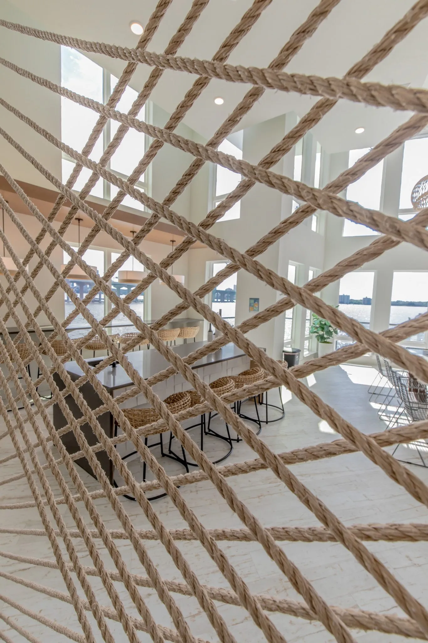 Multifamily amenity clubroom through a crisscross rope screen, woven stools at a long table in the double-height space