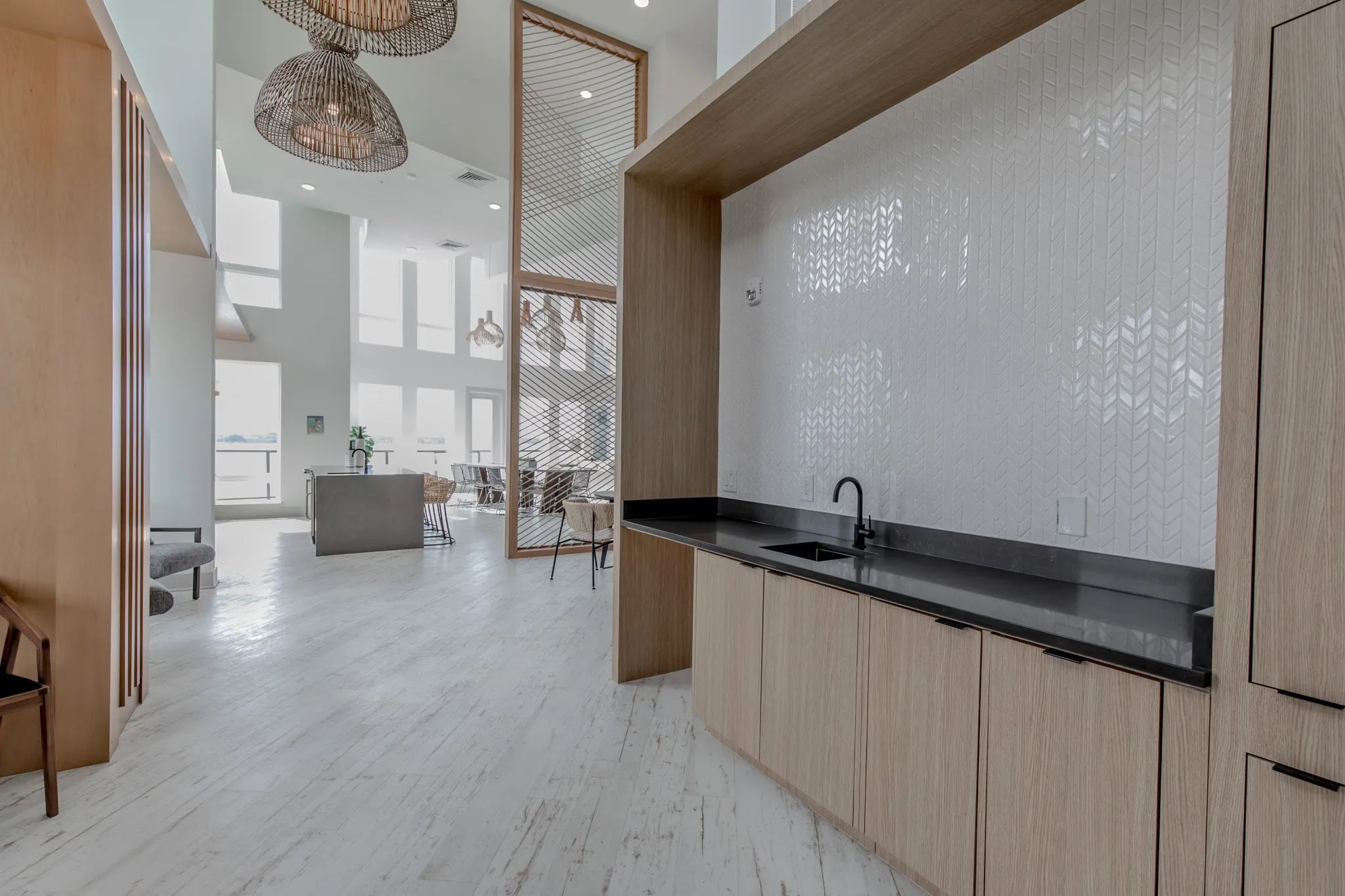 Multifamily amenity kitchenette with light oak cabinets and a black counter against white chevron tile, the clubroom beyond