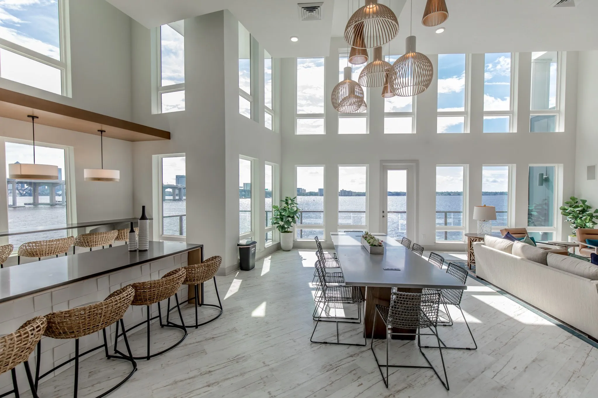 Multifamily amenity double-height clubroom with woven stools at a bar, a long table under wood pendants and a sofa