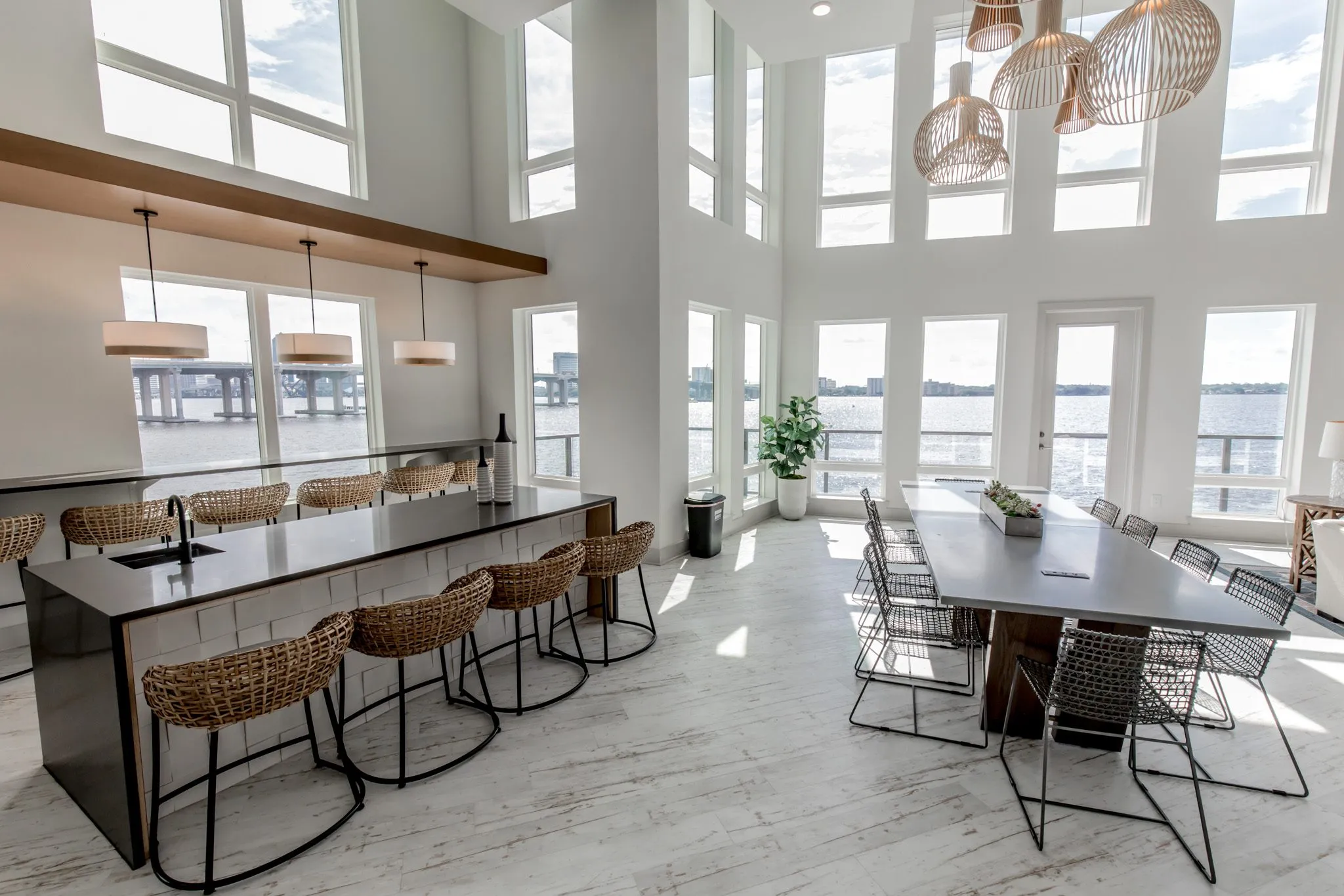 Multifamily amenity bar with woven stools and a long table with wire chairs under wood pendants, the river beyond