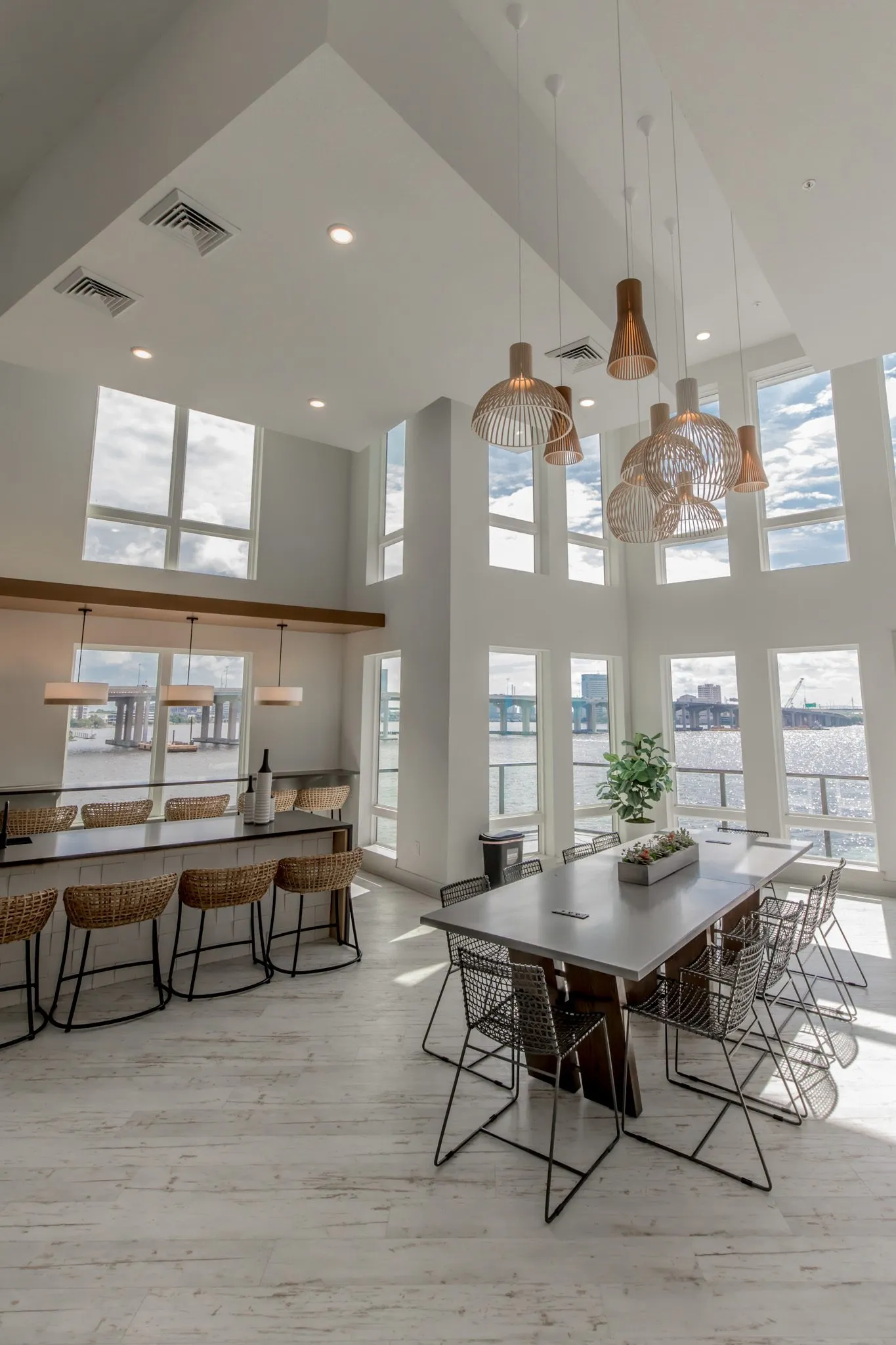 Multifamily amenity clubroom with a long table and wire chairs under clustered wood pendants, the bar and river windows