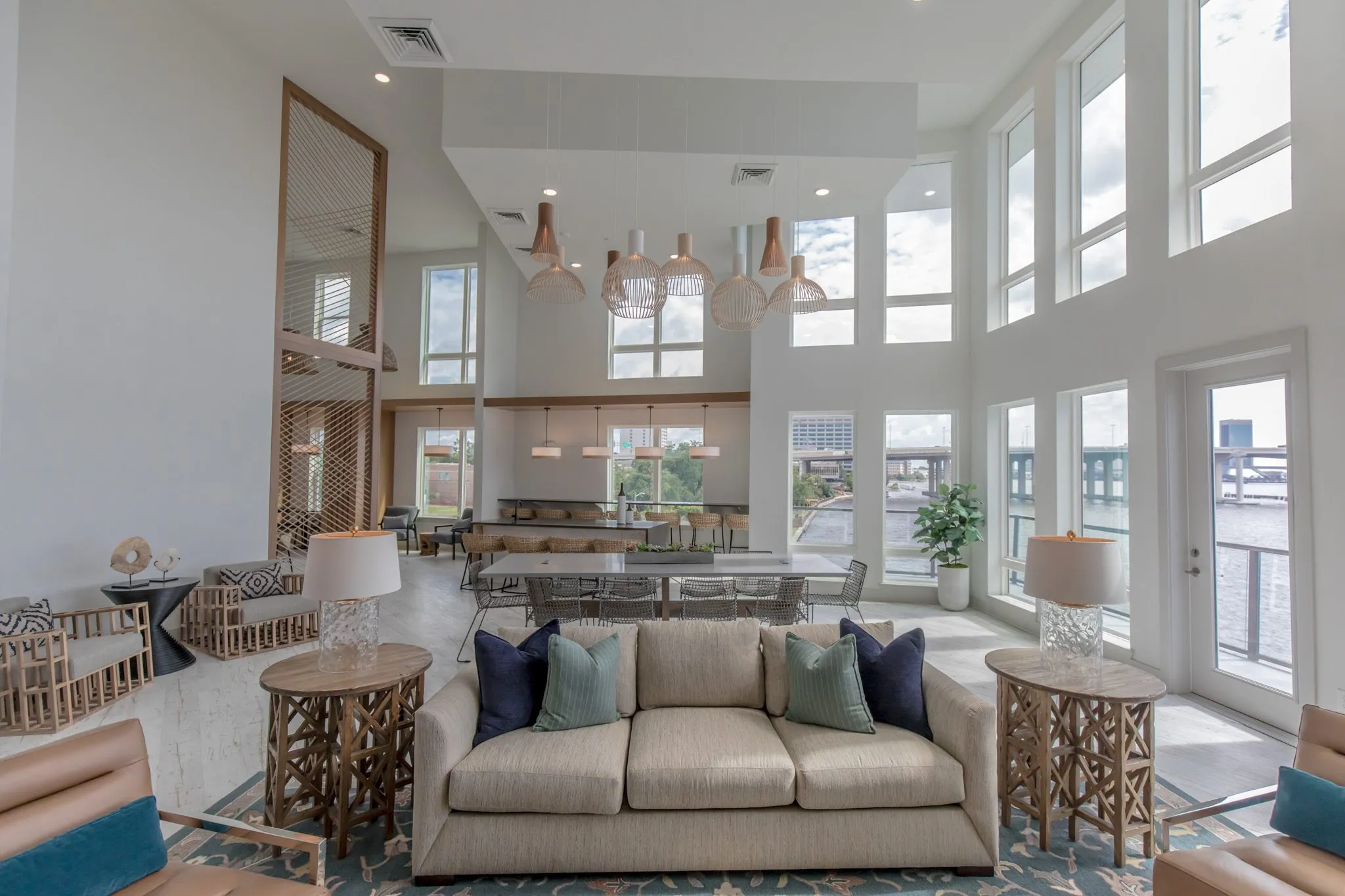 Multifamily amenity clubroom with a sofa and navy and green pillows between wood side tables and lamps, the long table beyond