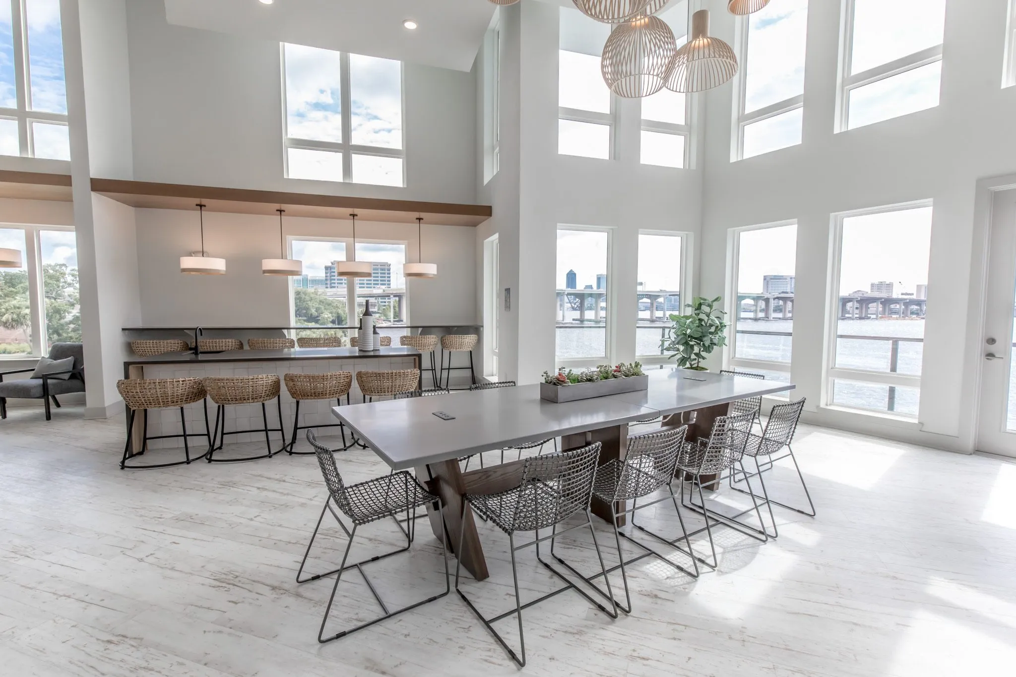 Multifamily amenity clubroom with a long table, wire chairs and a planter of succulents, the bar and river windows behind