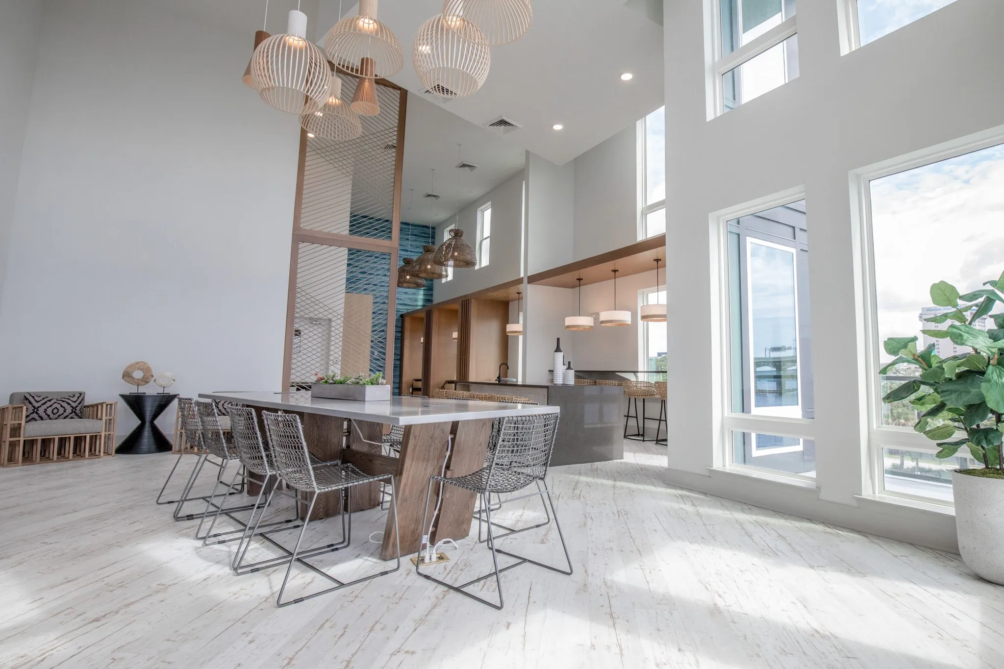 Multifamily amenity clubroom with the long table and wire chairs under wood pendants, a fiddle leaf fig by the windows
