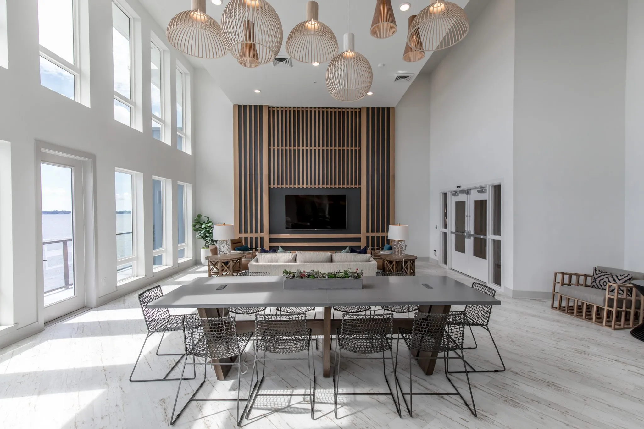 Multifamily amenity clubroom with a long table and wire chairs before the wood slat media wall, wood pendants overhead