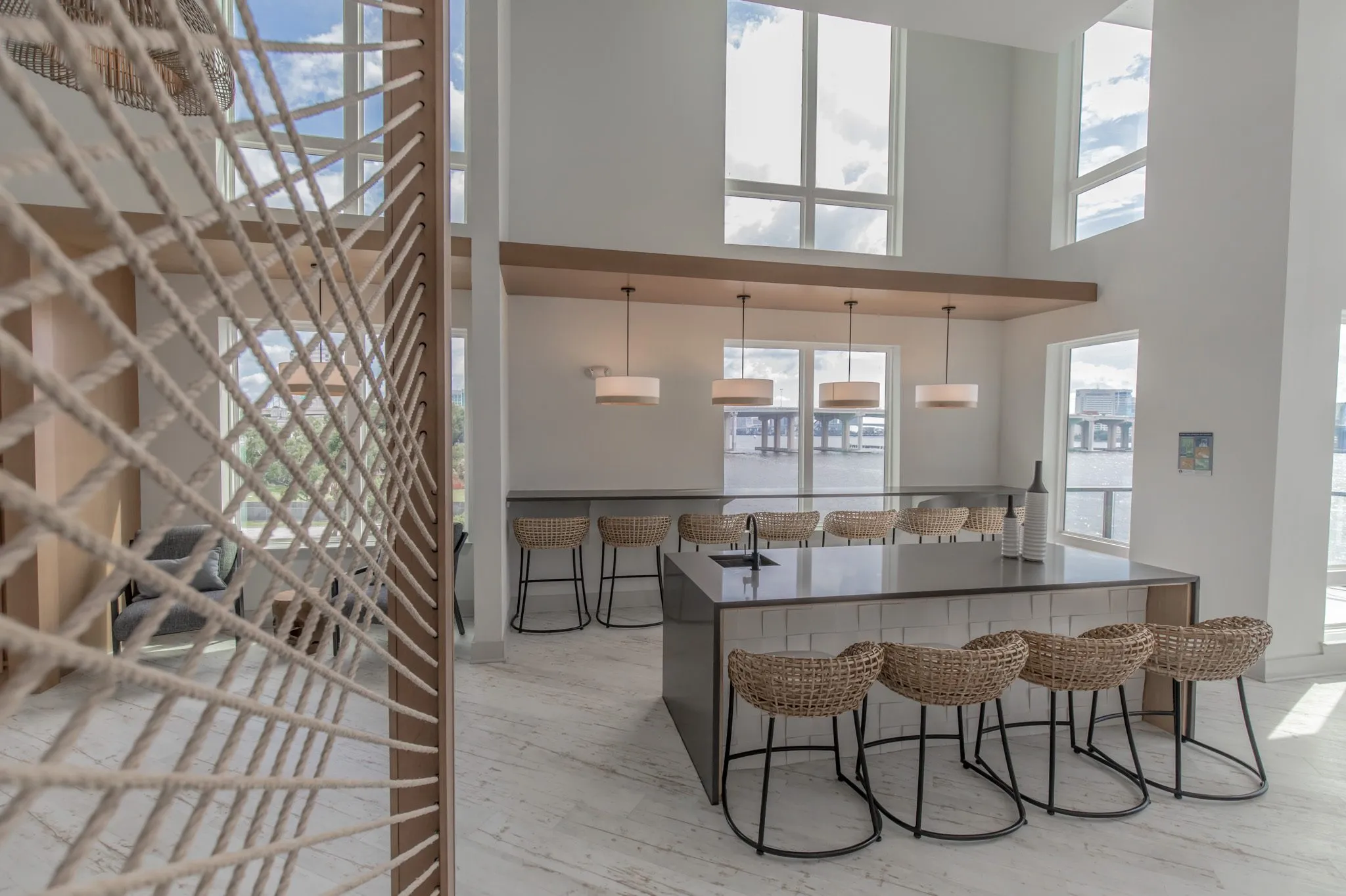 Multifamily amenity clubroom island with woven stools seen past a rope screen, a window bar under drum pendants