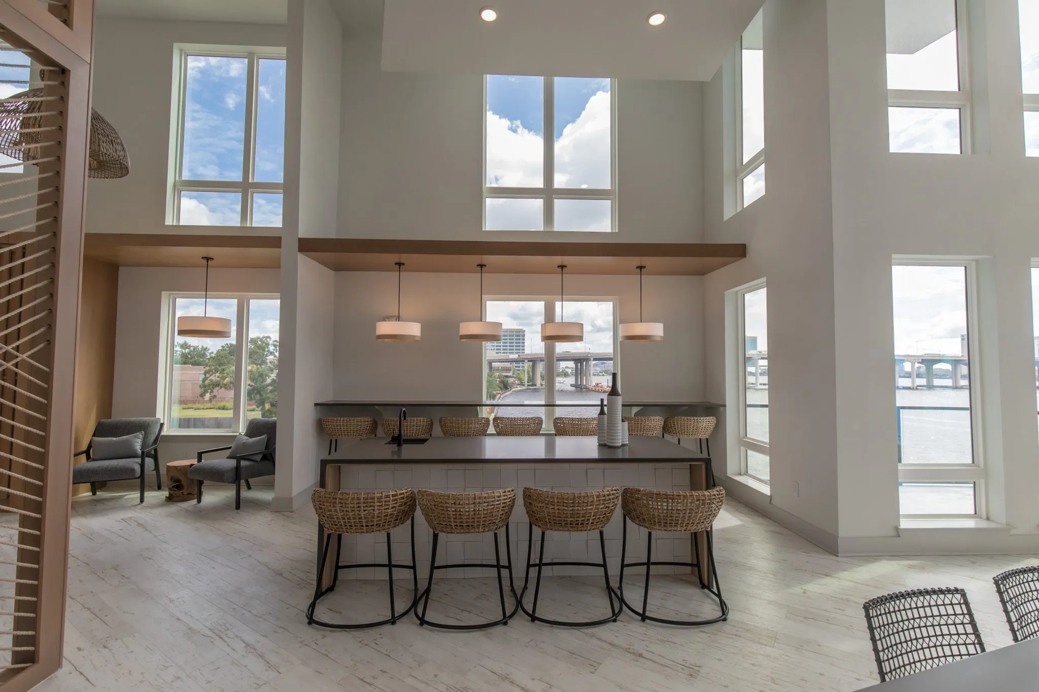 Multifamily amenity clubroom island with four woven stools before the window bar and drum pendants, the river beyond