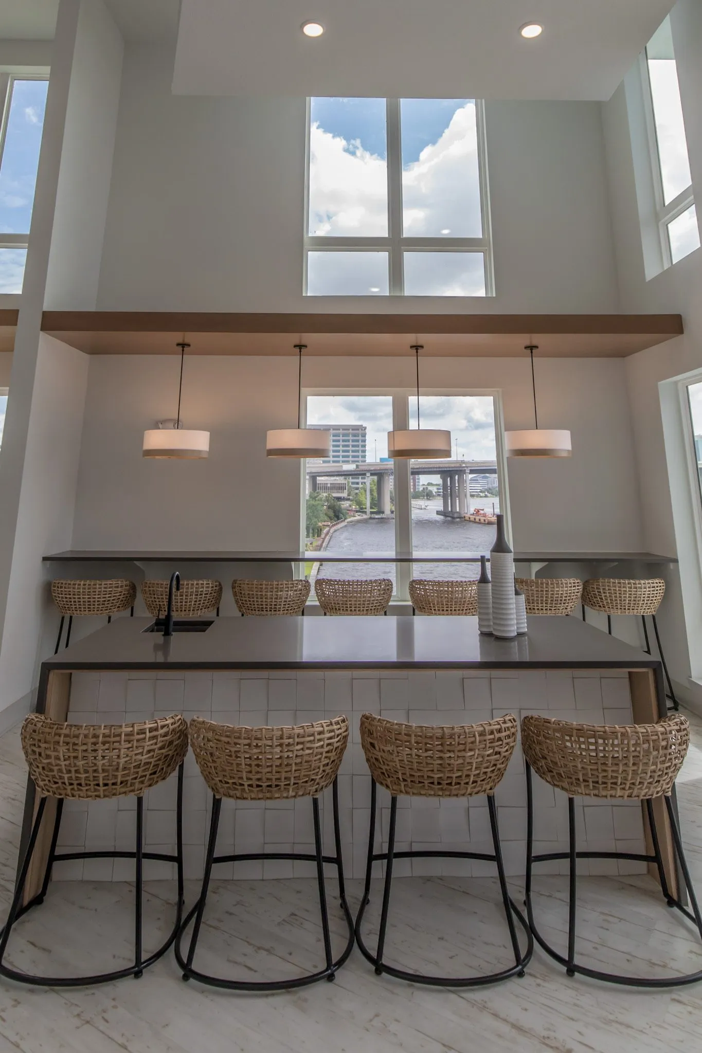 Multifamily amenity clubroom with woven stools at the island and window bar under drum pendants, the river beyond