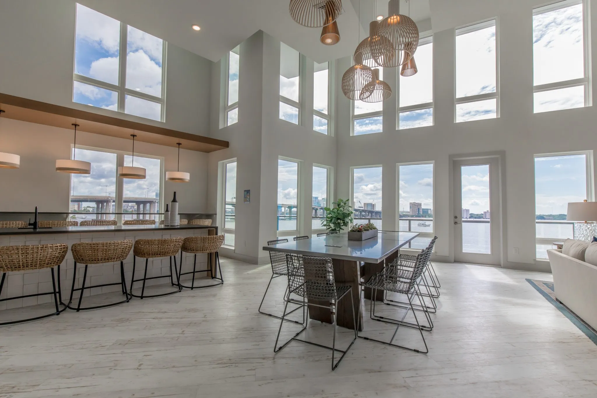 Multifamily amenity clubroom with woven stools at the island and a long table under clustered wood pendants, river windows