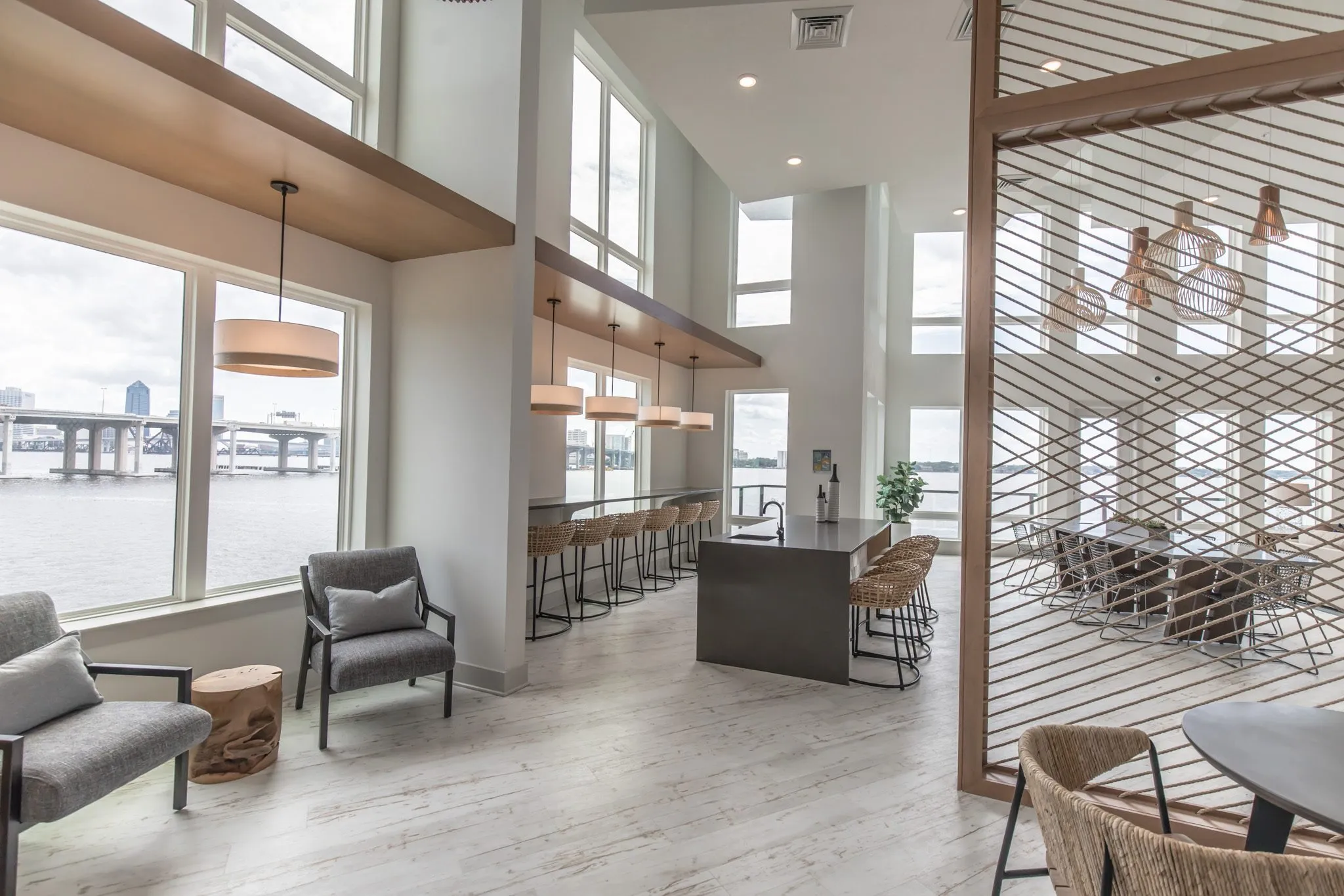 Multifamily amenity clubroom with grey armchairs by river windows near the bar and island, a rope screen at right