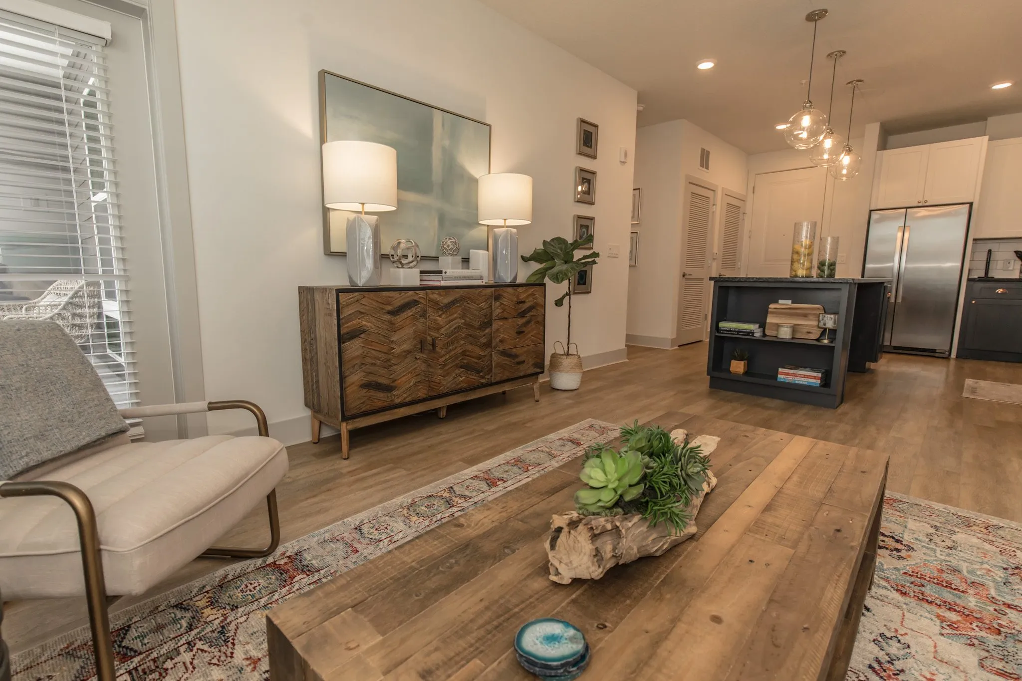Multifamily interior detail: wood coffee table with succulents on driftwood, a cream armchair, sideboard and kitchen beyond