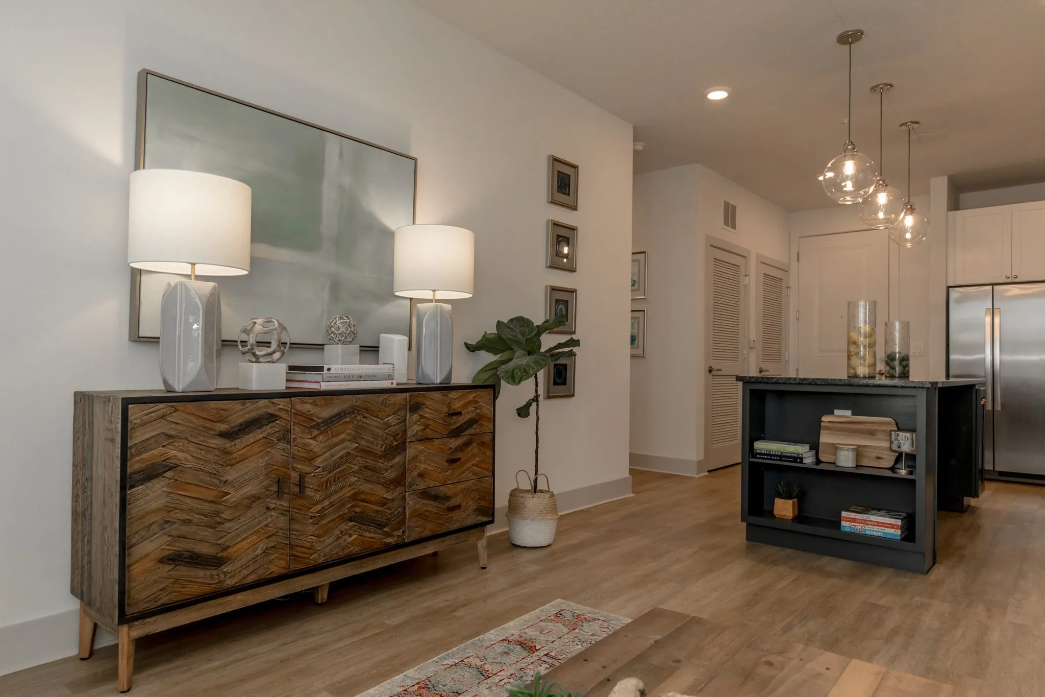 Multifamily interior detail: herringbone wood sideboard with white lamps below a large abstract painting, kitchen beyond