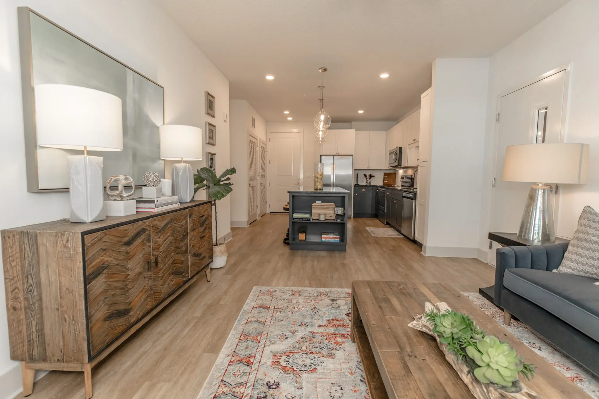 Multifamily apartment living room, past the sideboard and navy sofa to the kitchen with glass pendants and steel appliances
