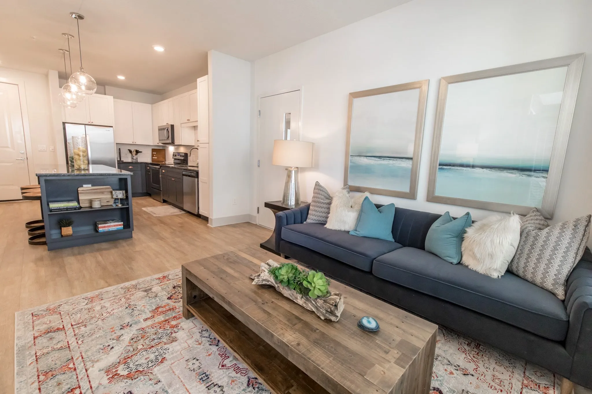 Multifamily apartment living room with a navy sofa below two framed seascapes, a wood coffee table and the kitchen at left