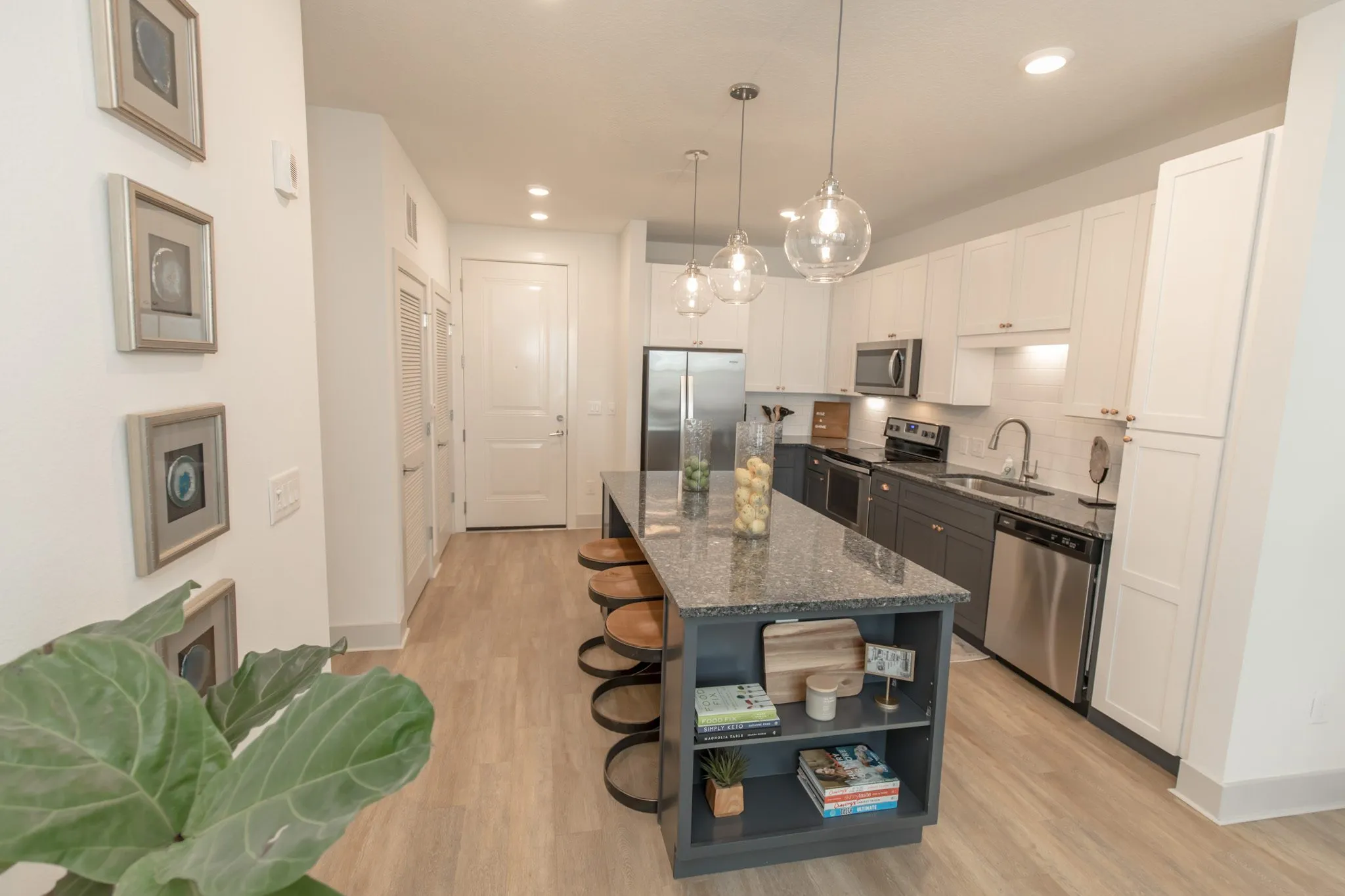 Multifamily kitchen seen past a fig leaf and framed prints, island stools under glass pendants