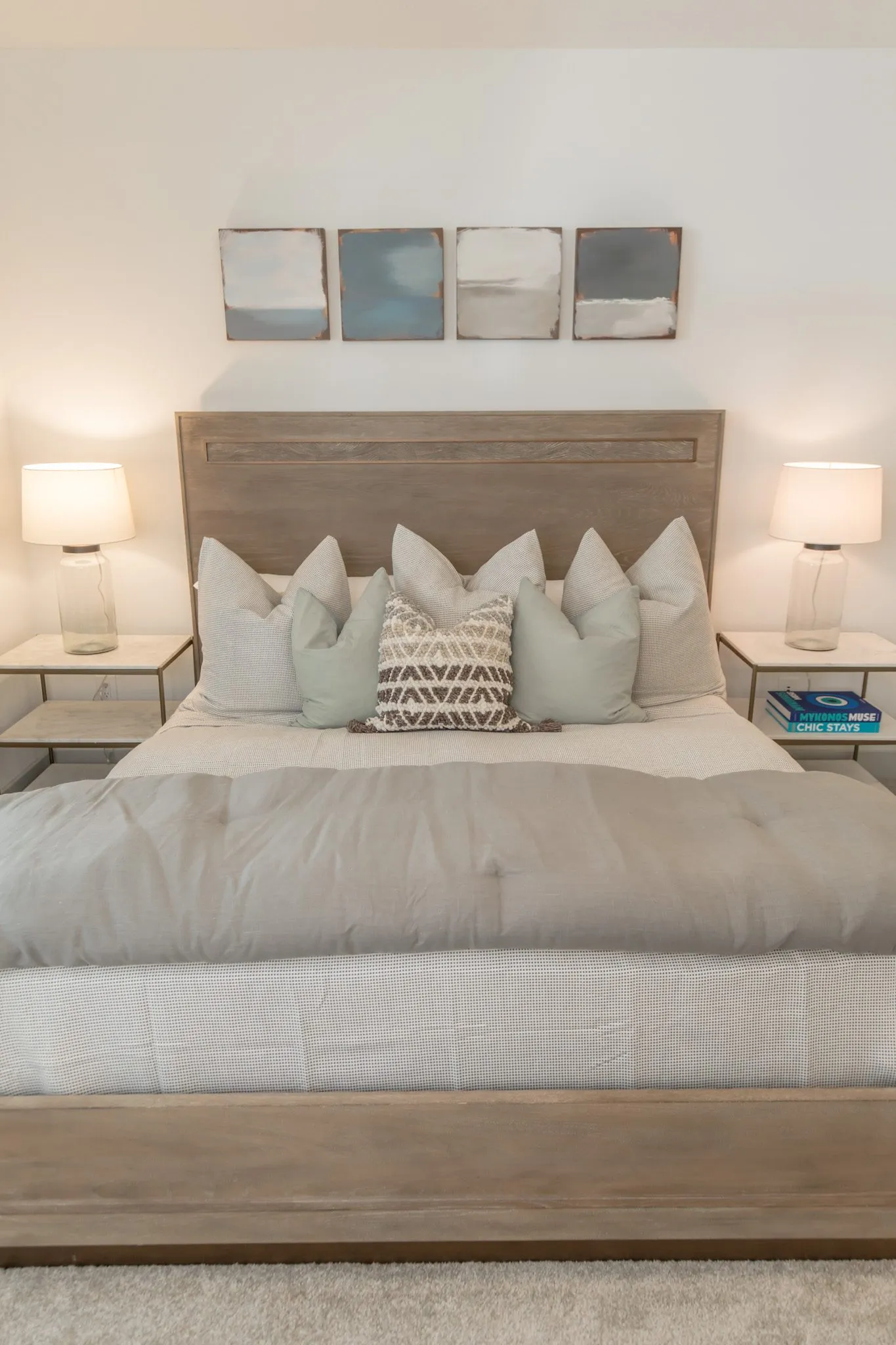Multifamily apartment bedroom with grey and sage pillows between glass lamps, four abstract prints above