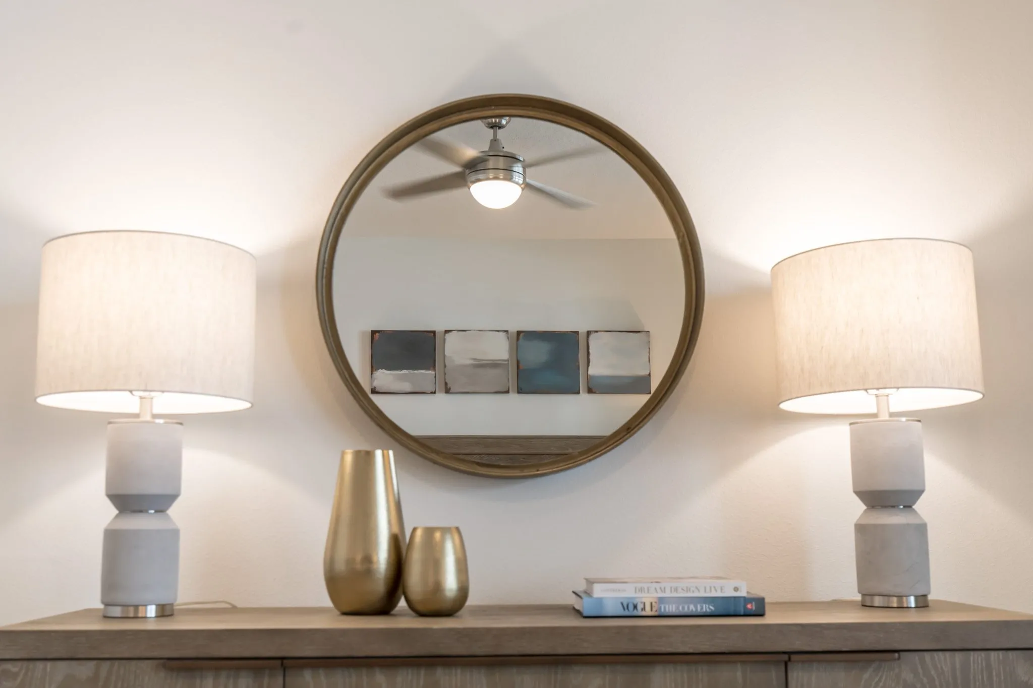 Multifamily interior detail: round wood mirror between two ceramic lamps above a dresser with brass vases and books