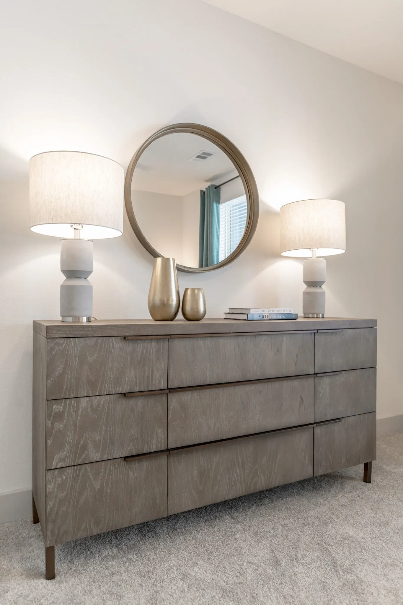 Multifamily interior detail: grey wood dresser with ceramic lamps and brass vases below a round mirror reflecting curtains