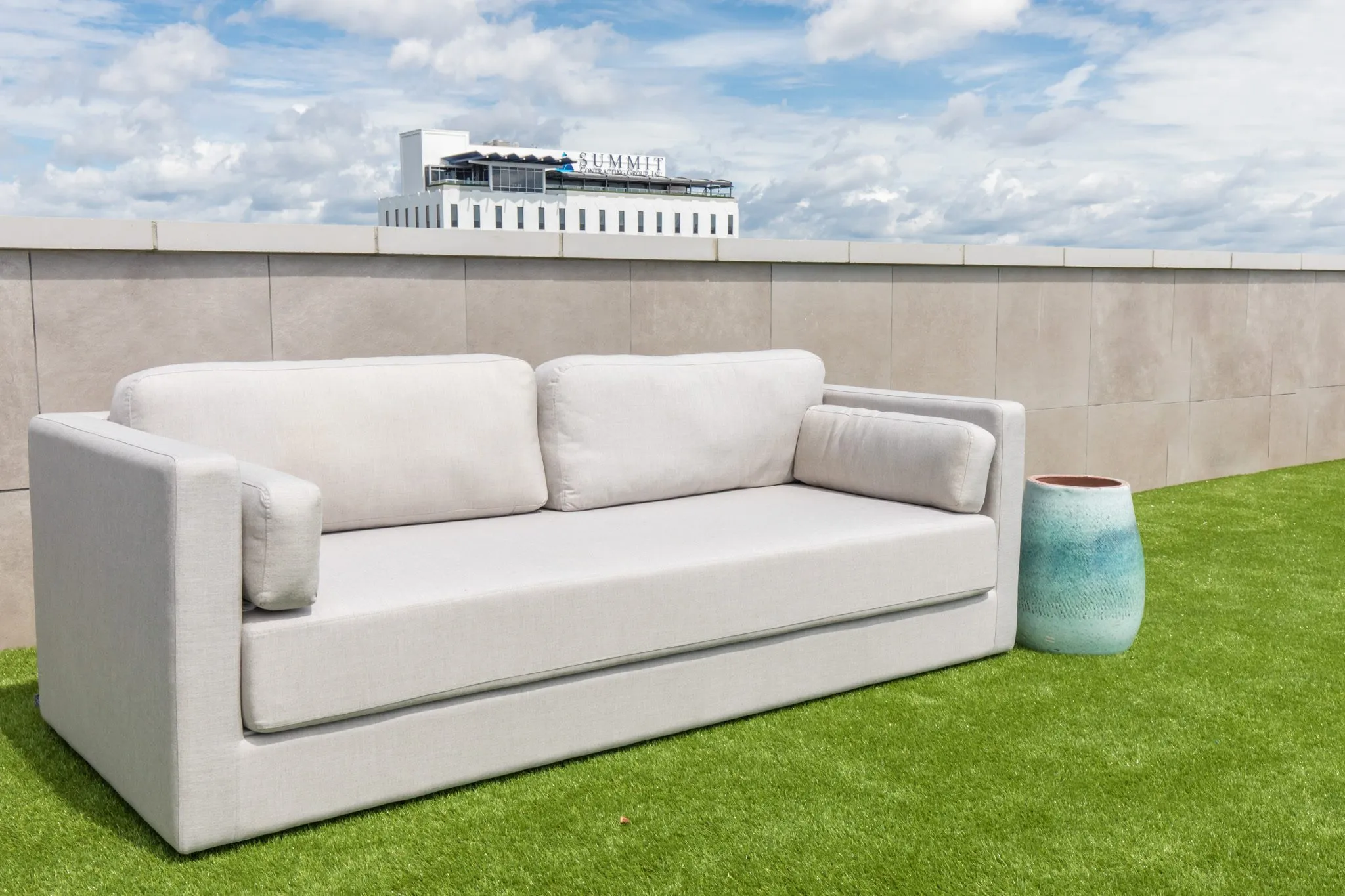 Multifamily outdoor seating: light grey sofa and a teal planter on artificial turf before a stone wall, a building beyond