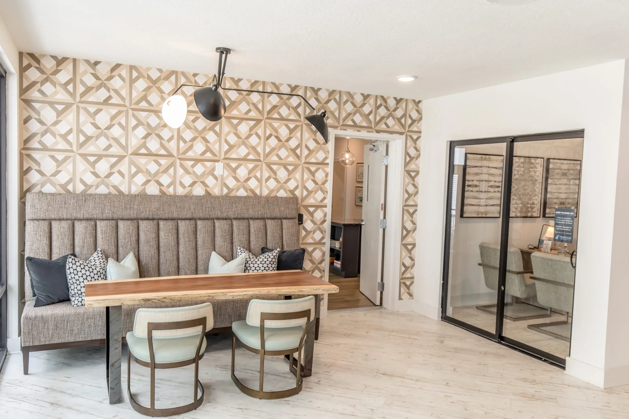 Multifamily amenity leasing area with a banquette and live edge table below a black swing-arm lamp, an office behind glass