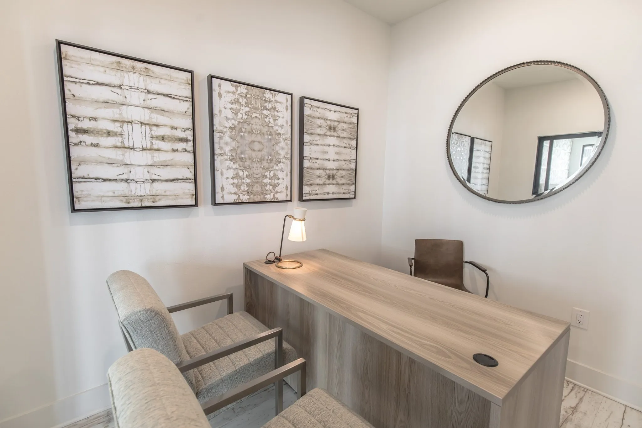 Multifamily amenity small office with a grey wood desk, two upholstered chairs, three textured prints and a round mirror