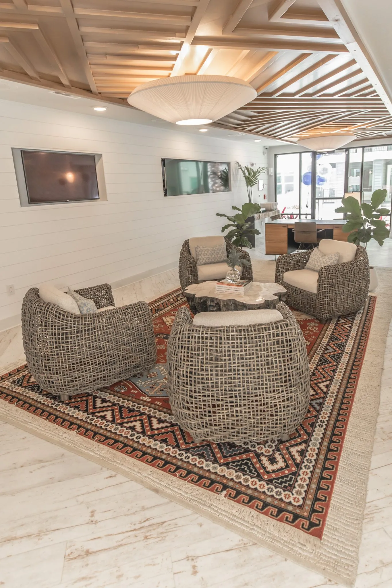 Multifamily amenity leasing lounge with woven armchairs around a stump table on a red patterned rug under a slatted ceiling