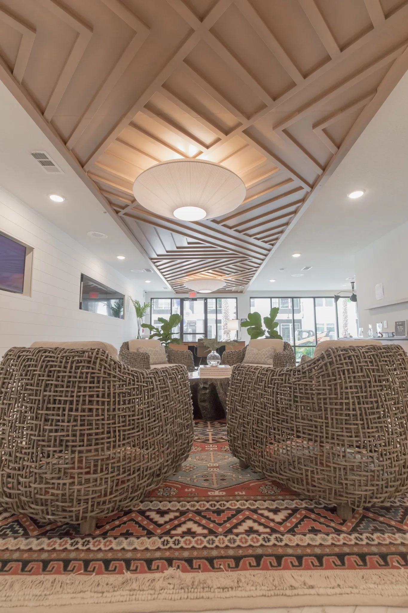 Multifamily amenity leasing lounge, a low view between woven armchairs down the rug to the windows under the slatted ceiling