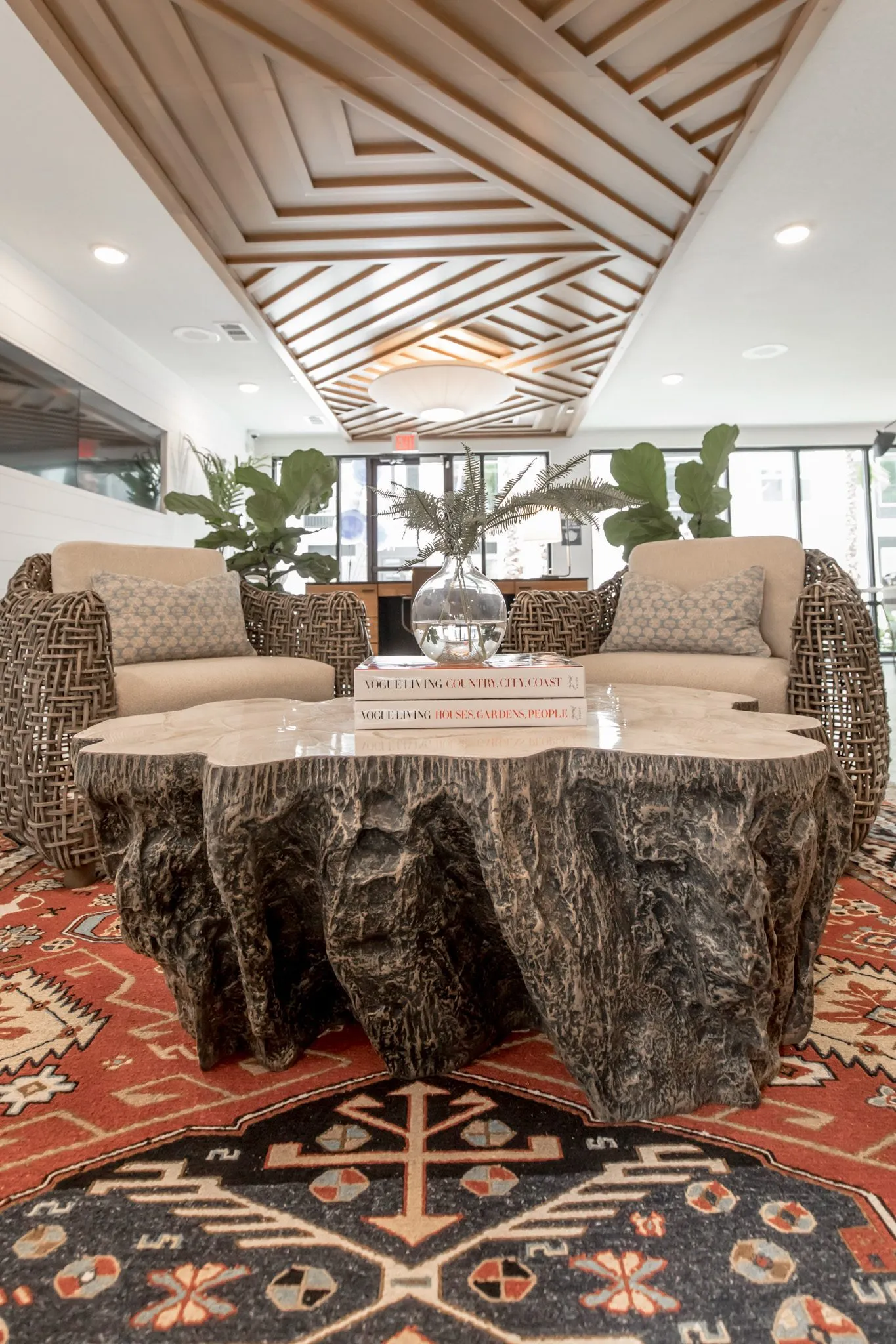 Multifamily interior detail: carved stump coffee table with a glass vase on books, woven armchairs and plants behind