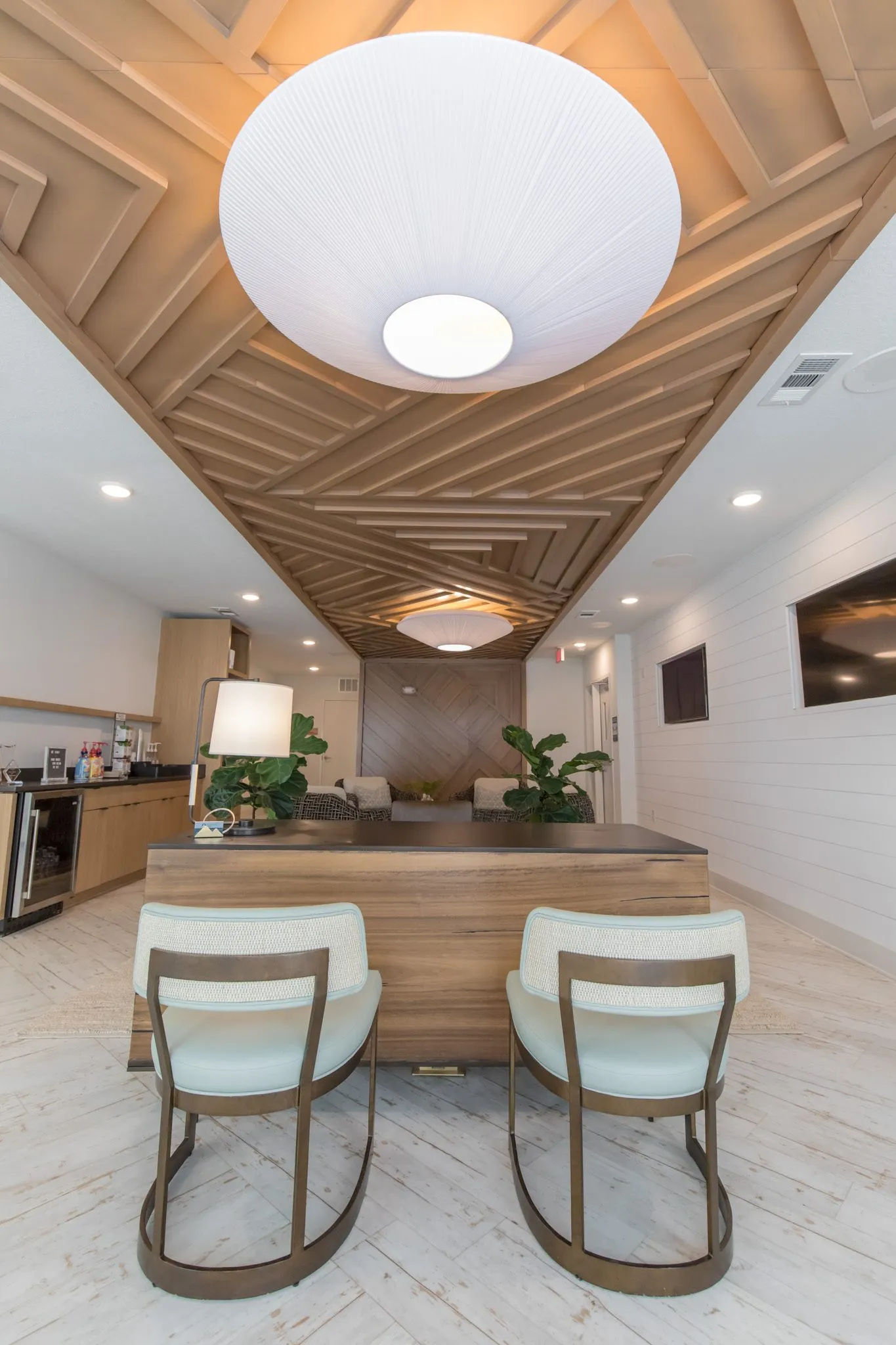 Multifamily amenity leasing desk with two sage chairs under a large pleated pendant, a lounge and herringbone wall beyond