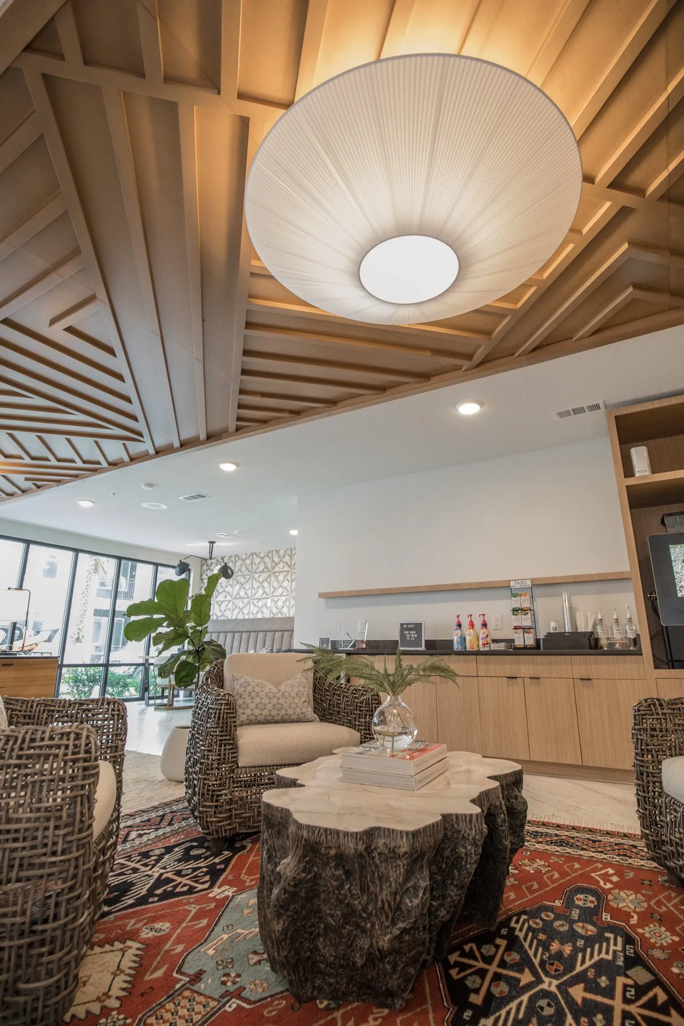 Multifamily amenity leasing lounge, woven armchairs around a stump table under a pleated pendant, oak cabinetry behind