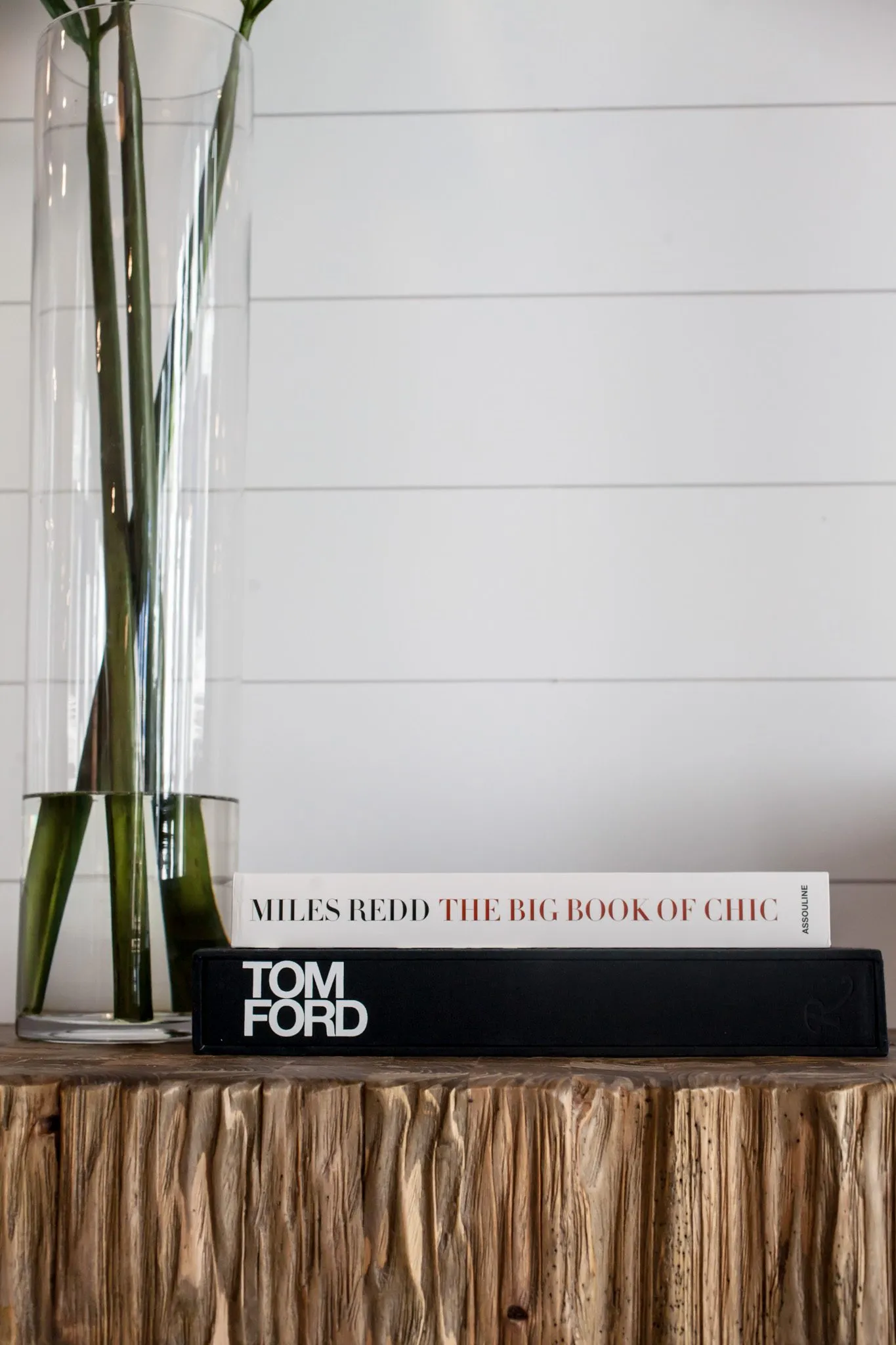 Multifamily interior detail: tall glass vase of green stems beside stacked books on a rough-hewn wood console by shiplap