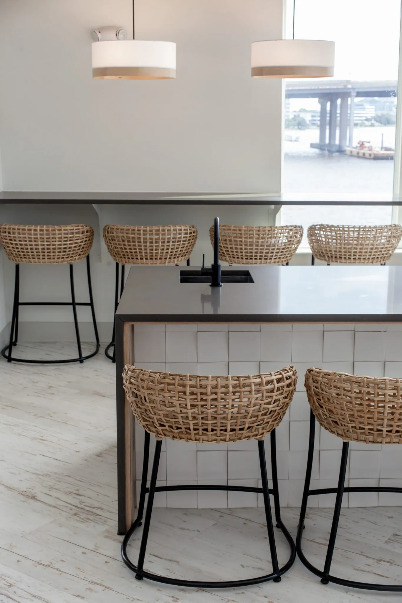 Multifamily amenity clubroom with woven stools at the island and window bar under drum pendants, the river and bridge beyond