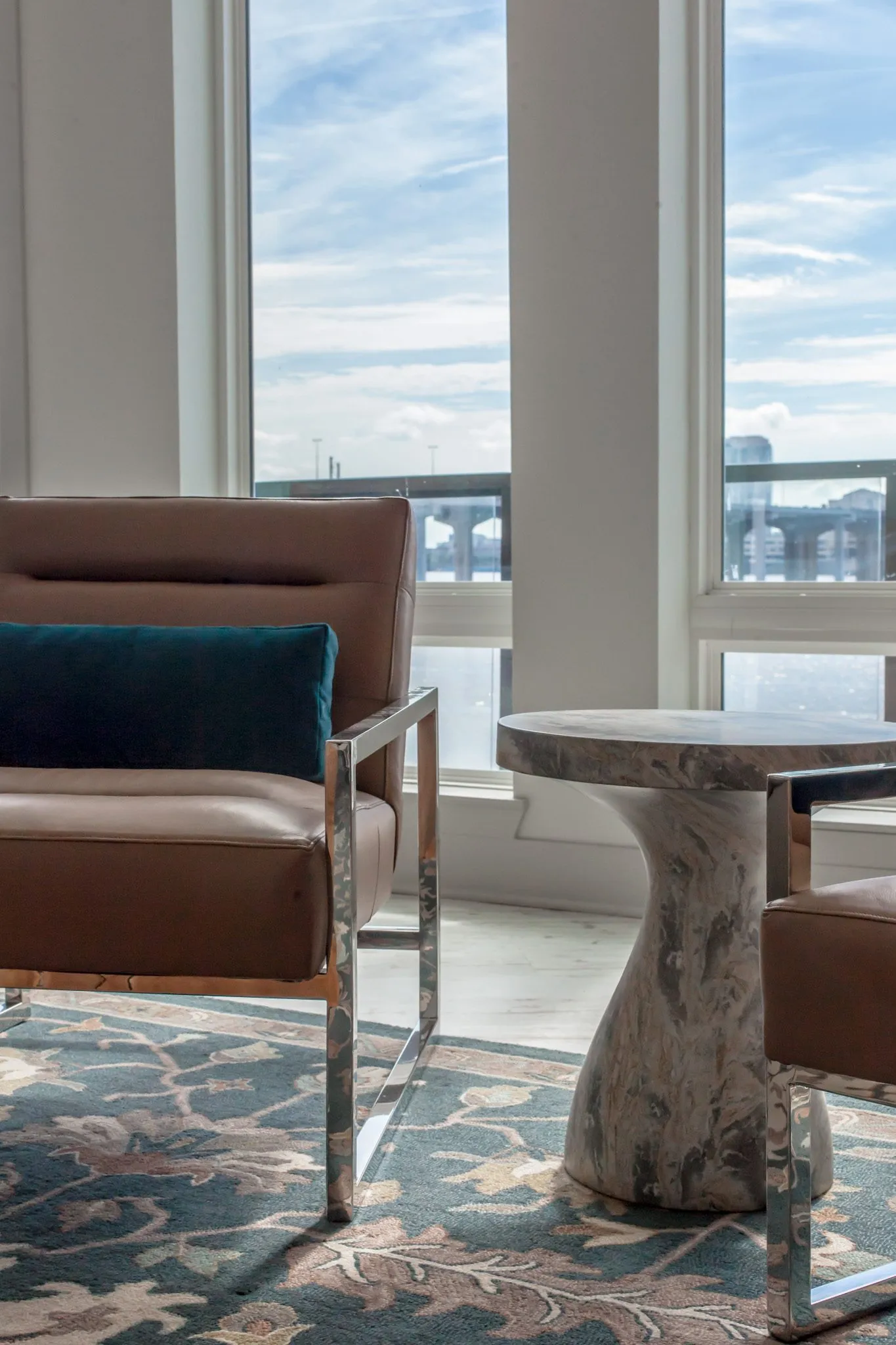 Multifamily interior detail: leather sling chair with a teal pillow beside a marble pedestal table by river windows