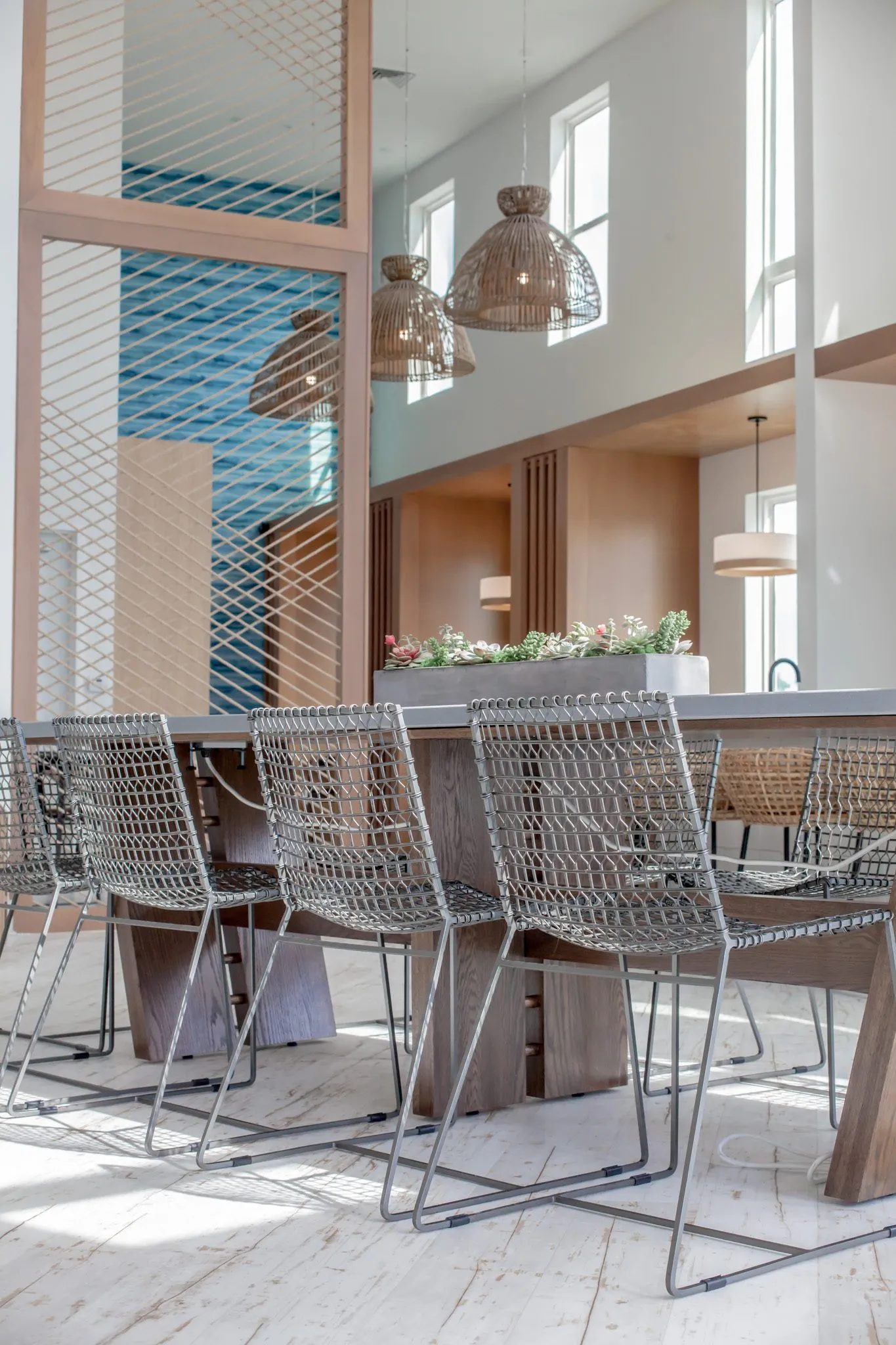 Multifamily amenity clubroom with wire chairs at the long table, the rope screen, woven pendants and booths beyond