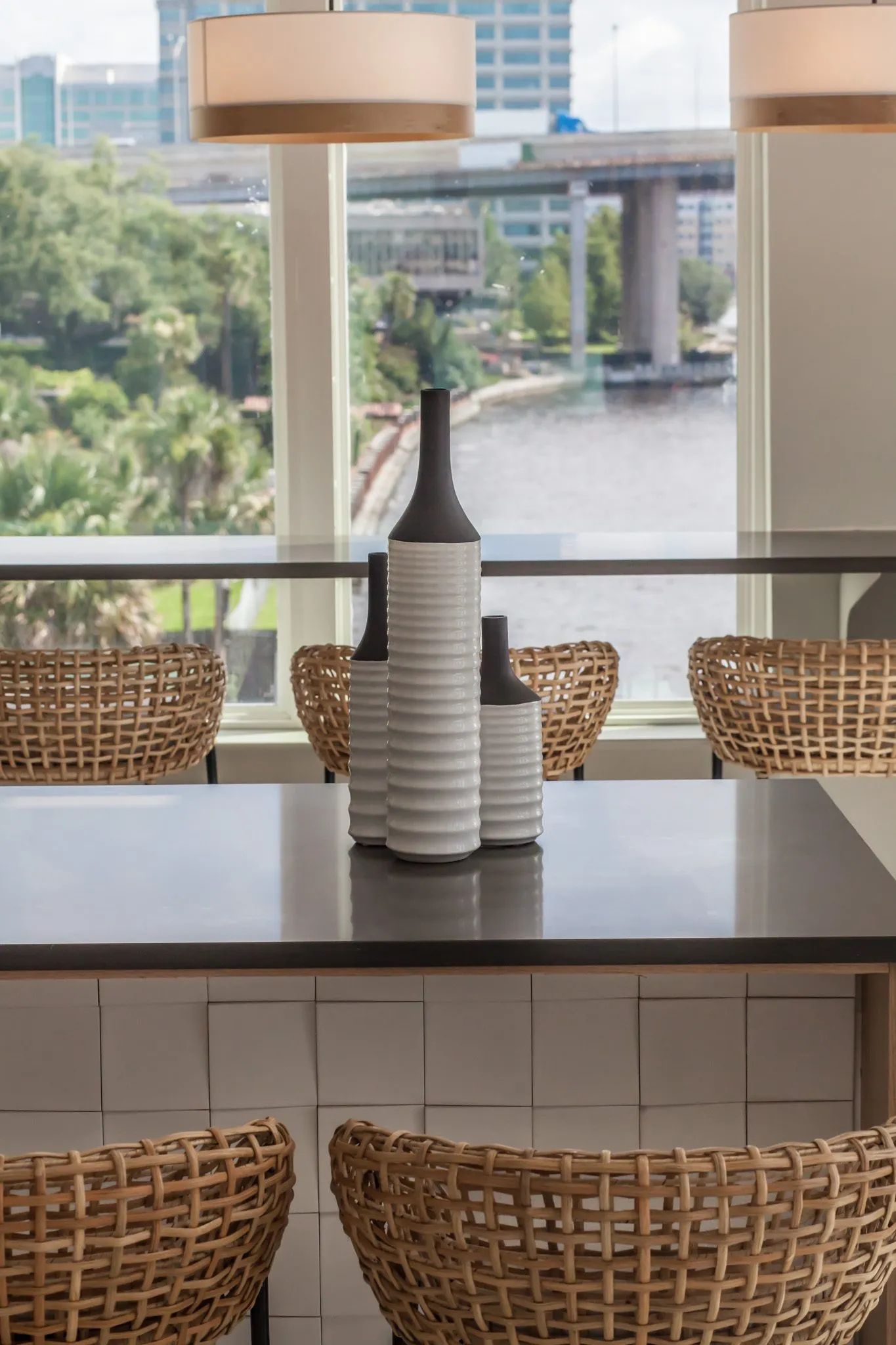 Multifamily interior detail: grey and white ceramic bottles on the island with woven stools, river and bridge beyond