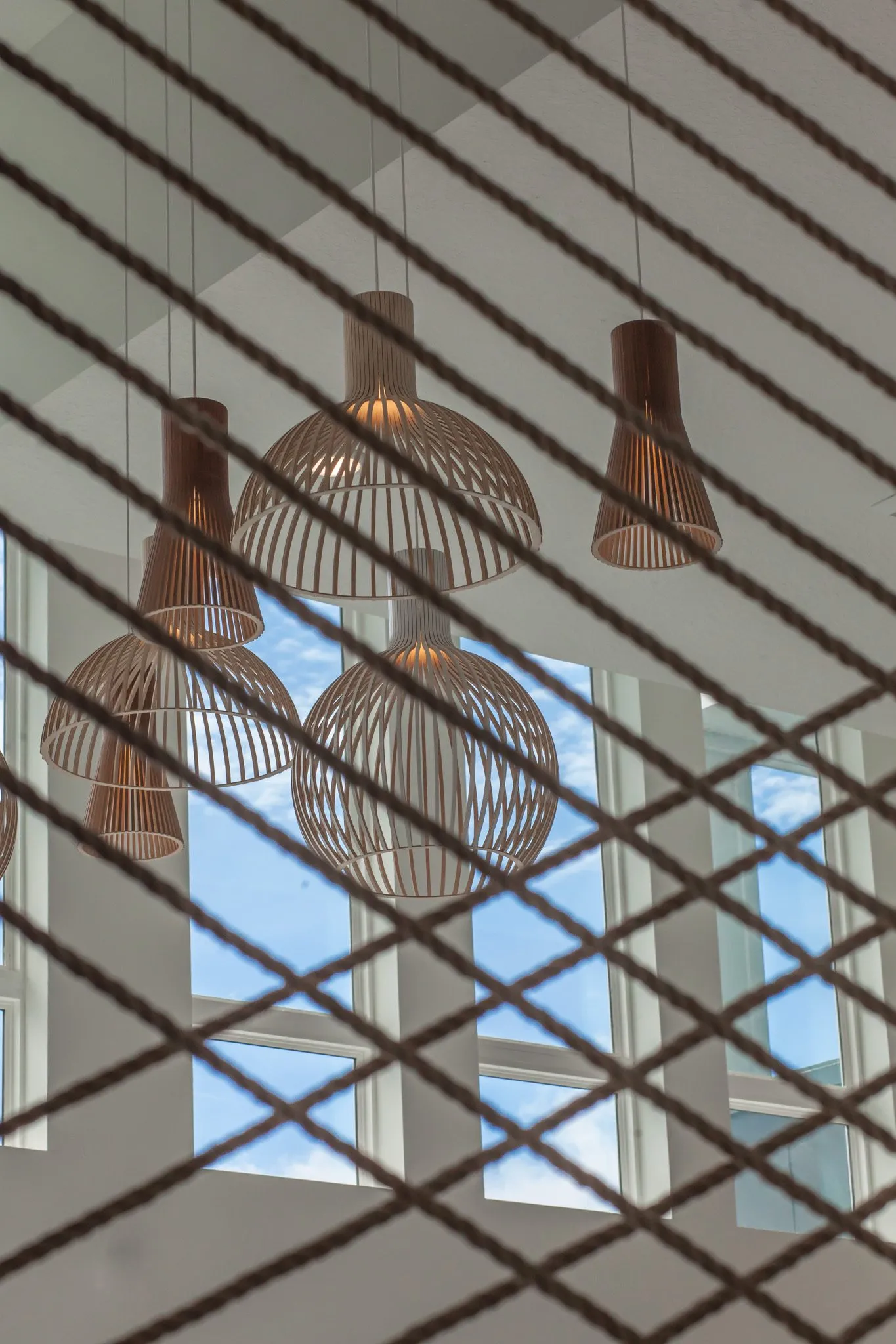Multifamily interior detail: wood ribbon pendants seen through the crisscross rope screen against tall windows