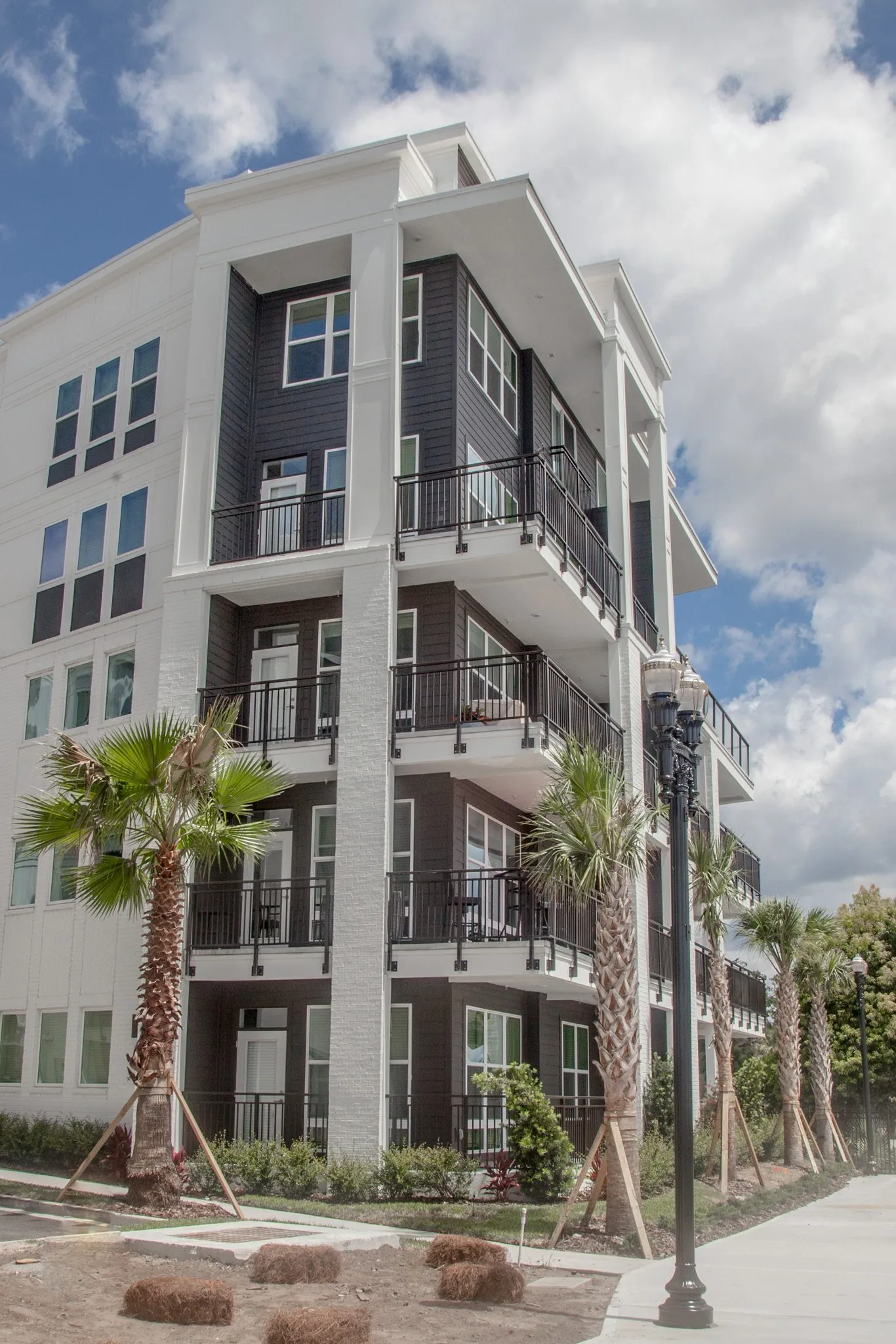 Multifamily community exterior: building corner with stacked balconies, a black lamp post and staked young palms